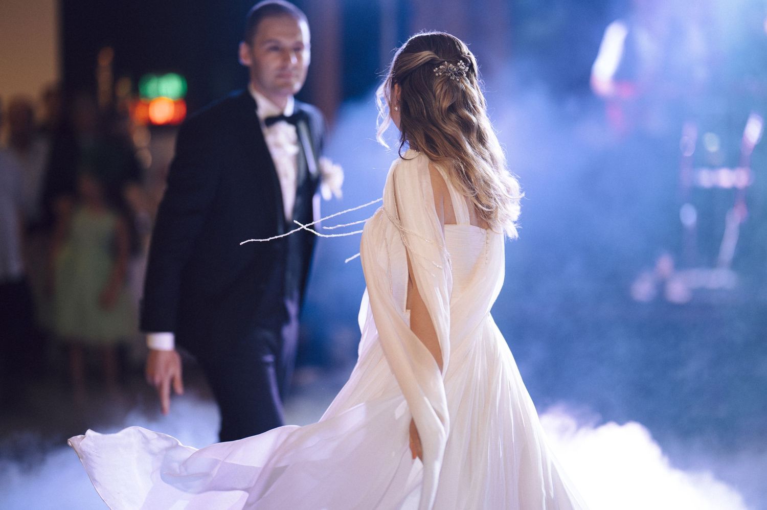 A bride in a flowing white dress spins during a romantic wedding first dance amid atmospheric lighting and fog.