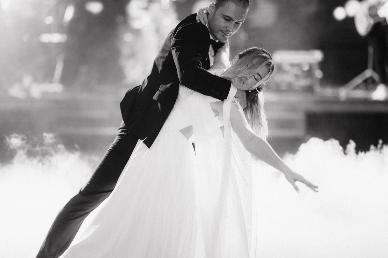 A graceful bride in a white gown and groom in black suit dance together surrounded by fog on their wedding day.