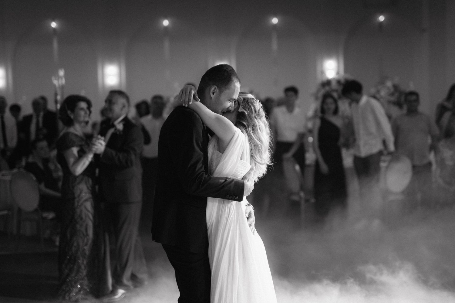 A couple shares their first dance as newlyweds on a misty dance floor while guests watch in black and white.