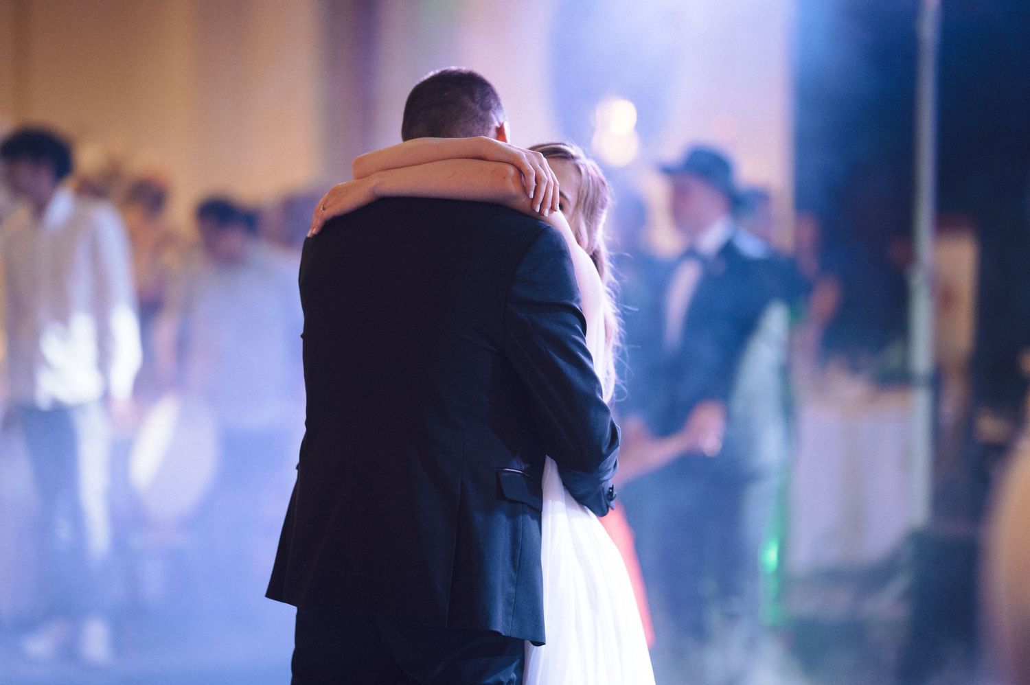 A couple shares an intimate dance at what appears to be their wedding reception as guests look on in soft lighting.
