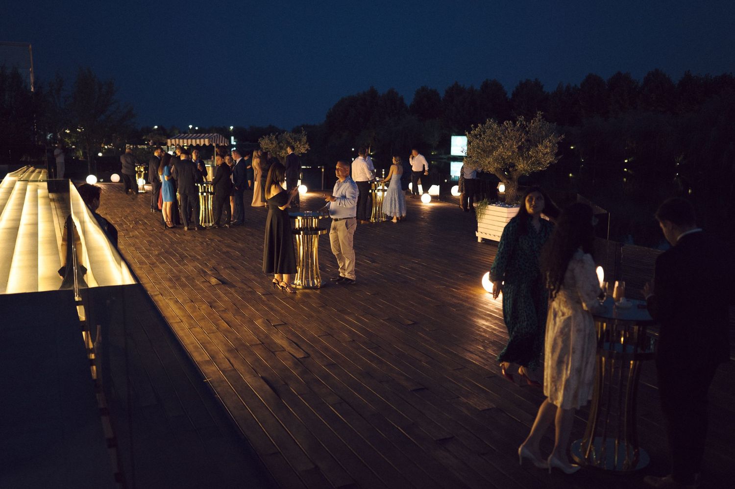 People gather on a wooden pier at night with glowing lights illuminating the evening waterfront scene.