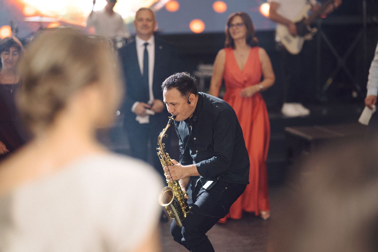 A saxophone player performs on a dance floor at an evening event with people in formal attire visible in the background.