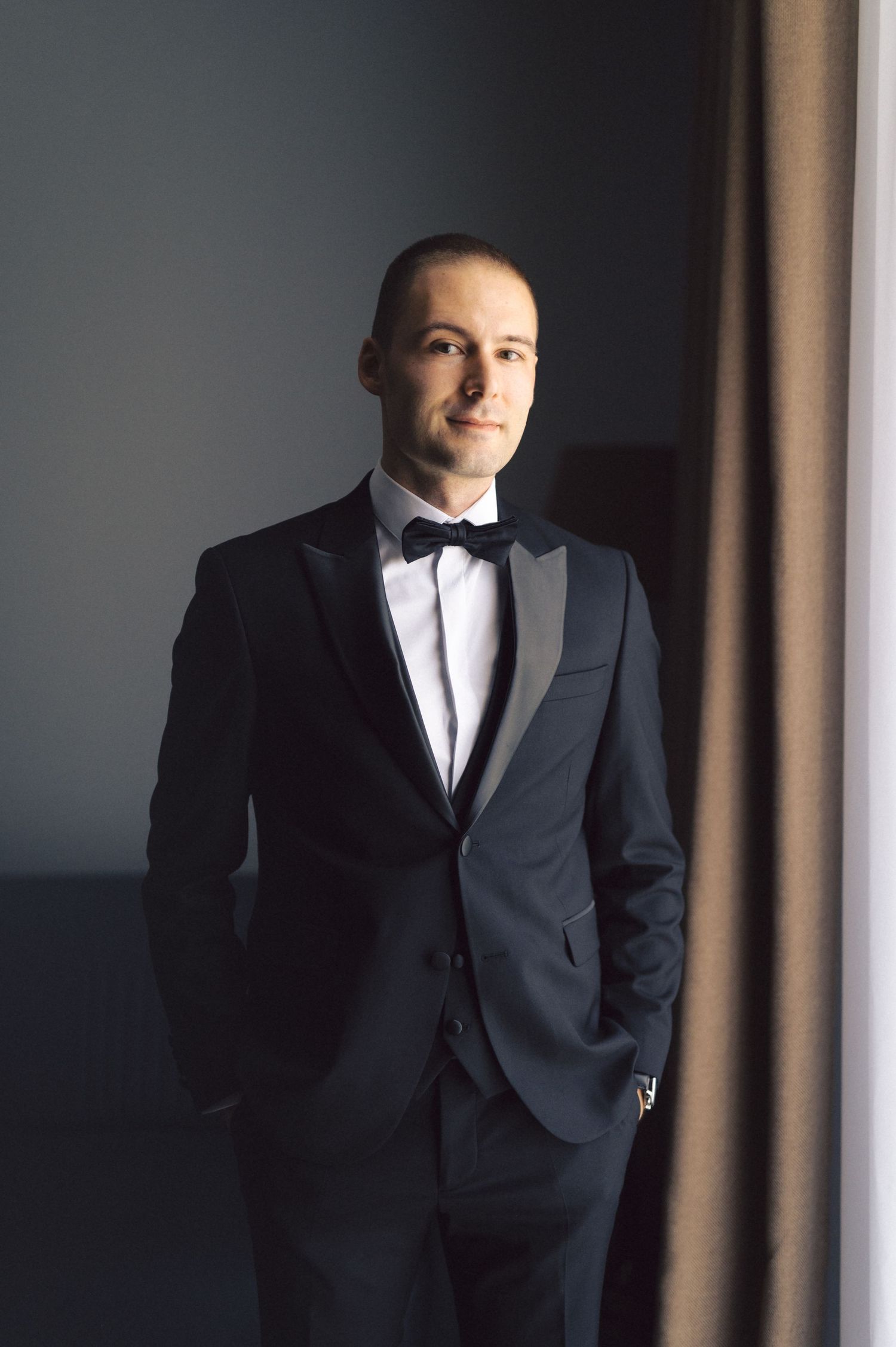 A person in a classic black tuxedo with bow tie poses near a window in moody natural lighting.