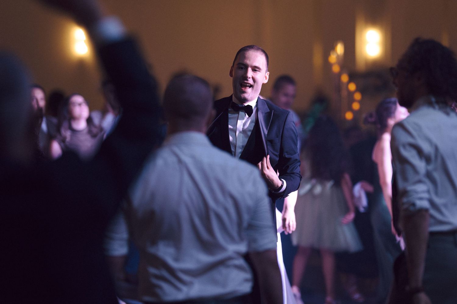 A performer speaks into a microphone on a dimly lit dance floor surrounded by party guests.