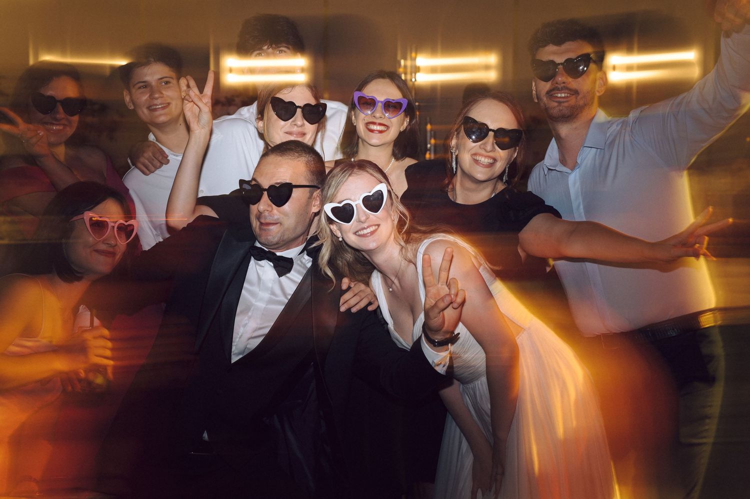Wedding party wearing sunglasses pose together in a dimly lit indoor setting during reception celebrations.