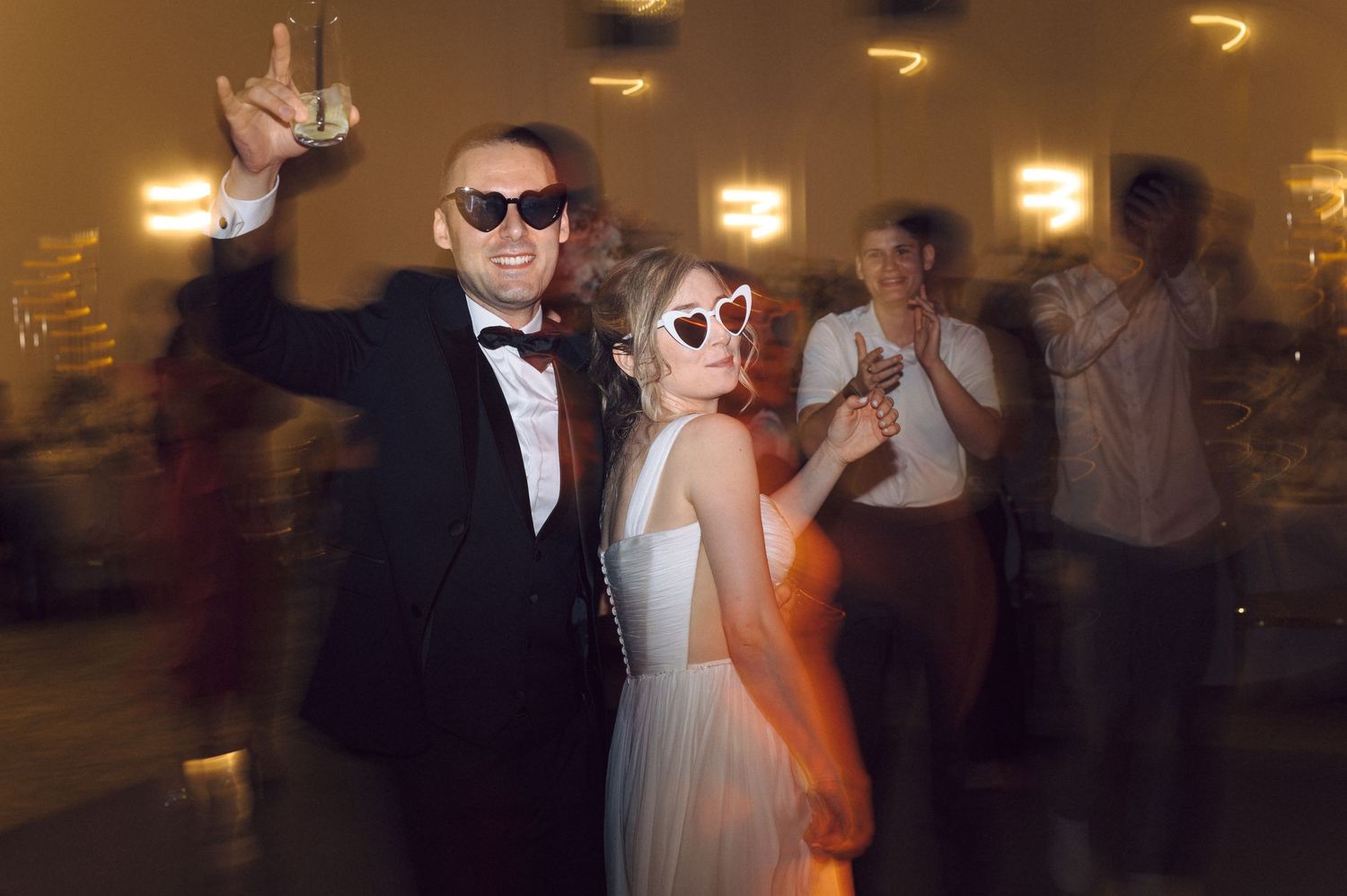 Wedding guests dance and celebrate in a dark reception hall with warm lighting and festive atmosphere.