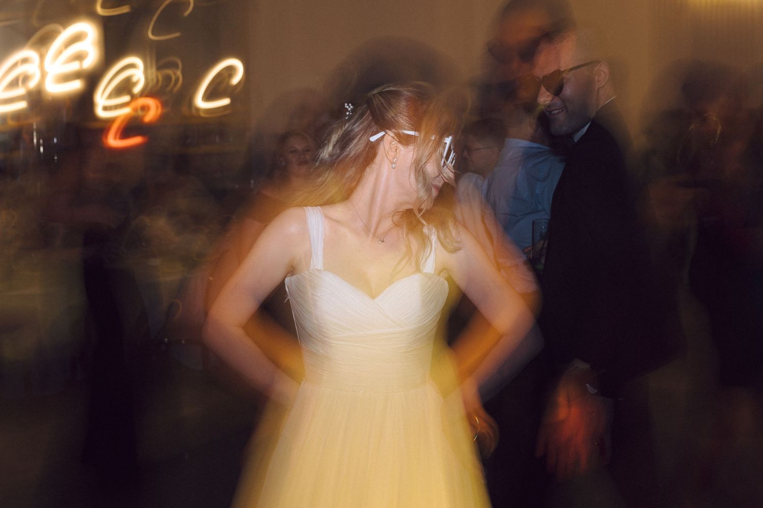 Motion-blurred photograph of figures dancing at a wedding reception, with a white dress in focus against blurry lights.