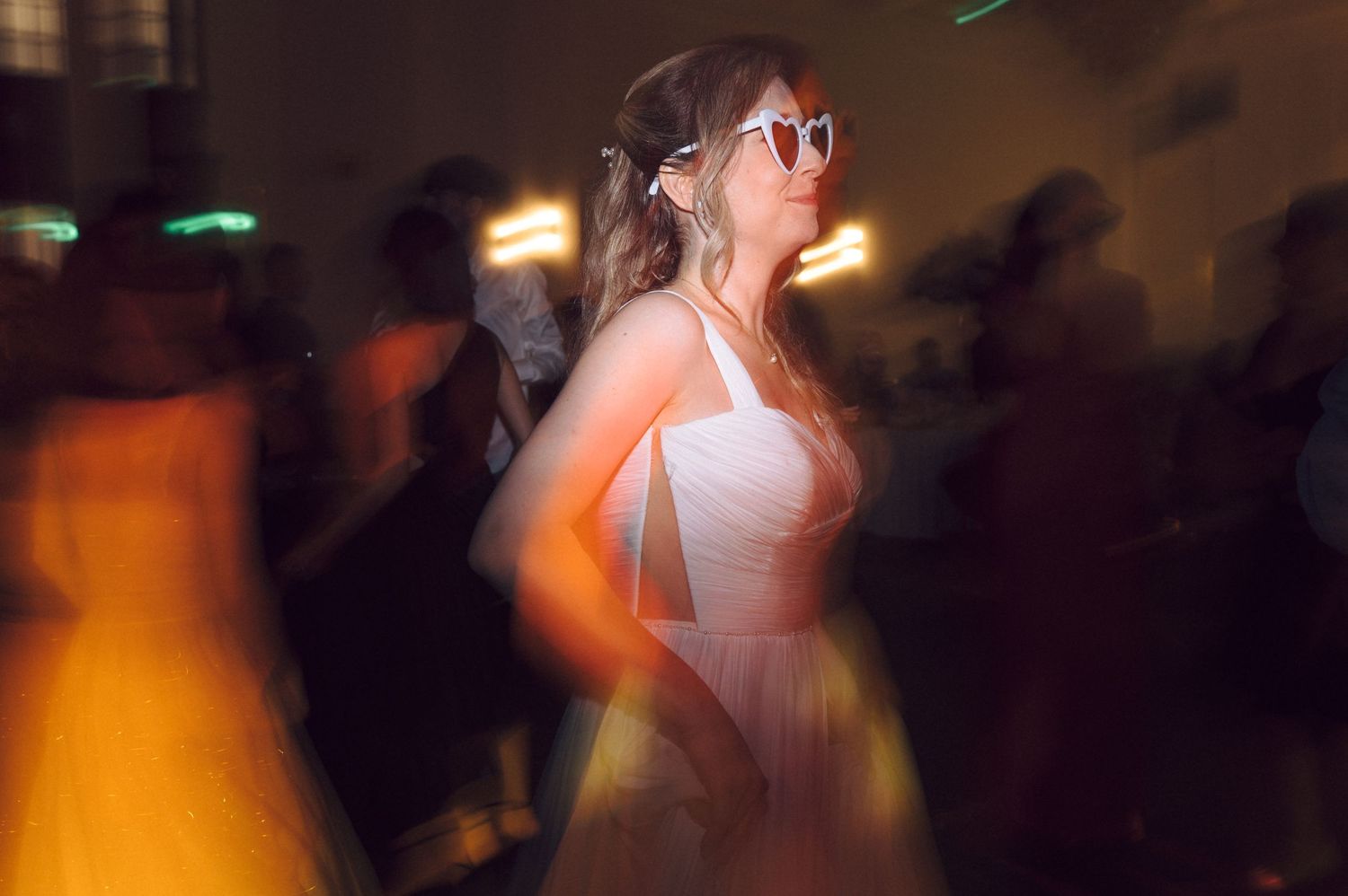 A bride in a white dress dancing at a dimly lit wedding reception with soft glowing lights in the background.
