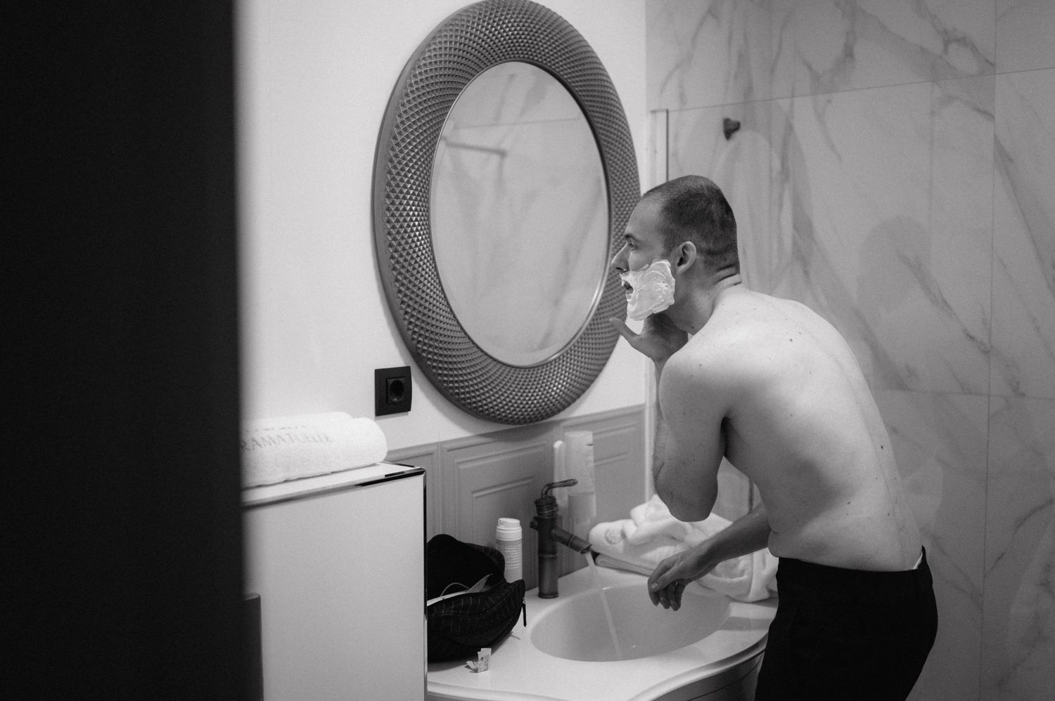 A shirtless person leans over a bathroom sink while looking into a circular mirror mounted on a marble wall.