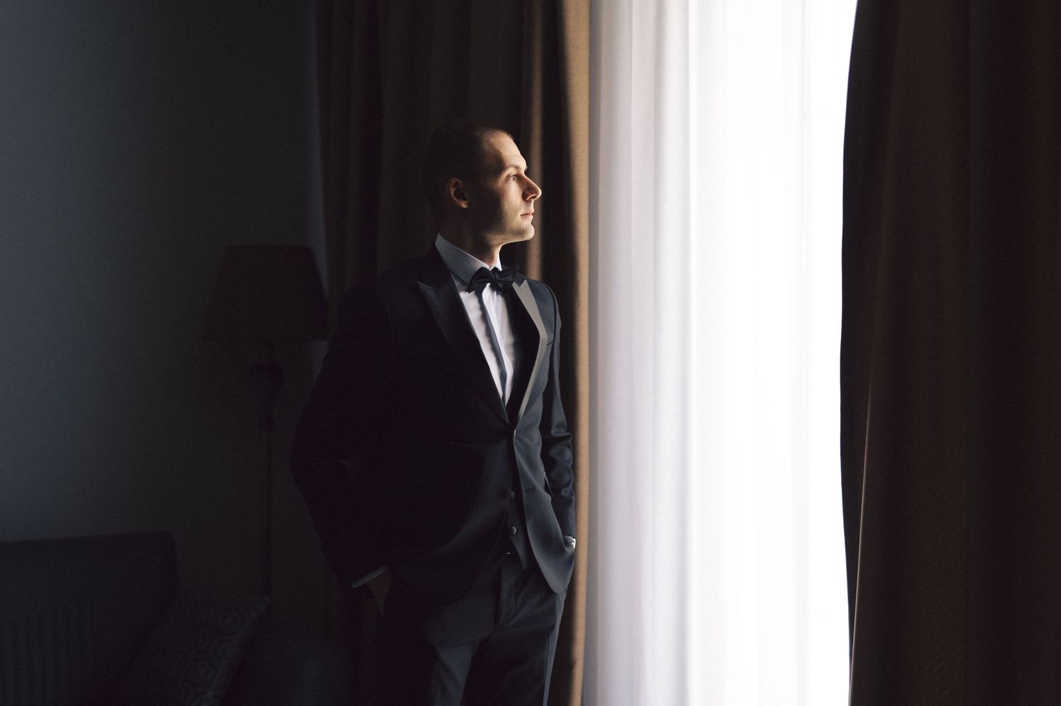 Professional silhouetted figure in business attire stands pensively by window in dramatic lighting.