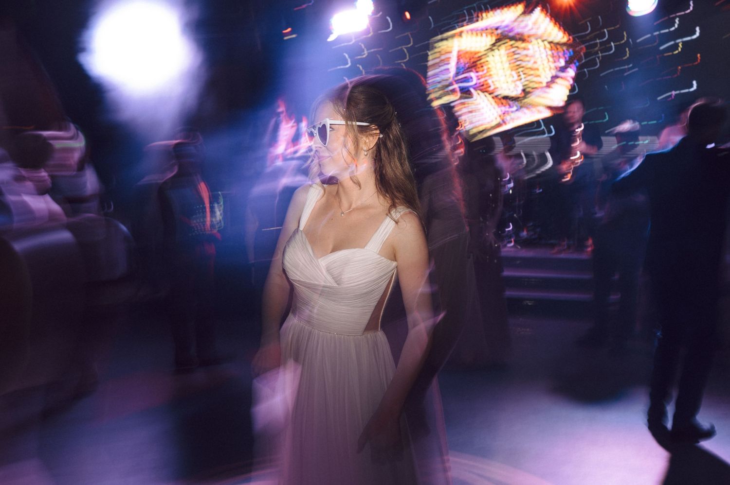Ethereal white dress glows under purple club lights as blurred party guests dance in the background.