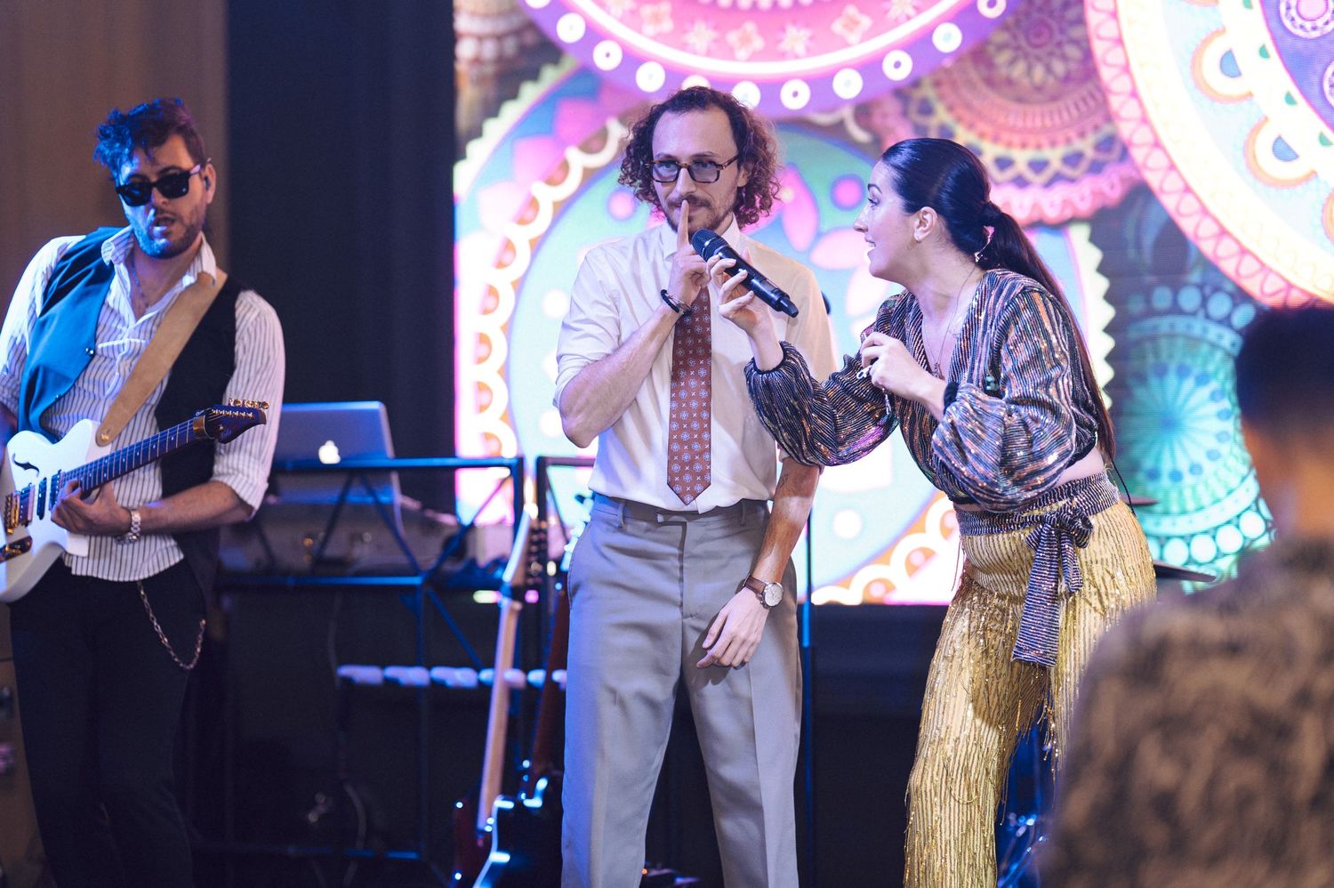 Musicians perform on stage with colorful mandala artwork projected on screen behind them.