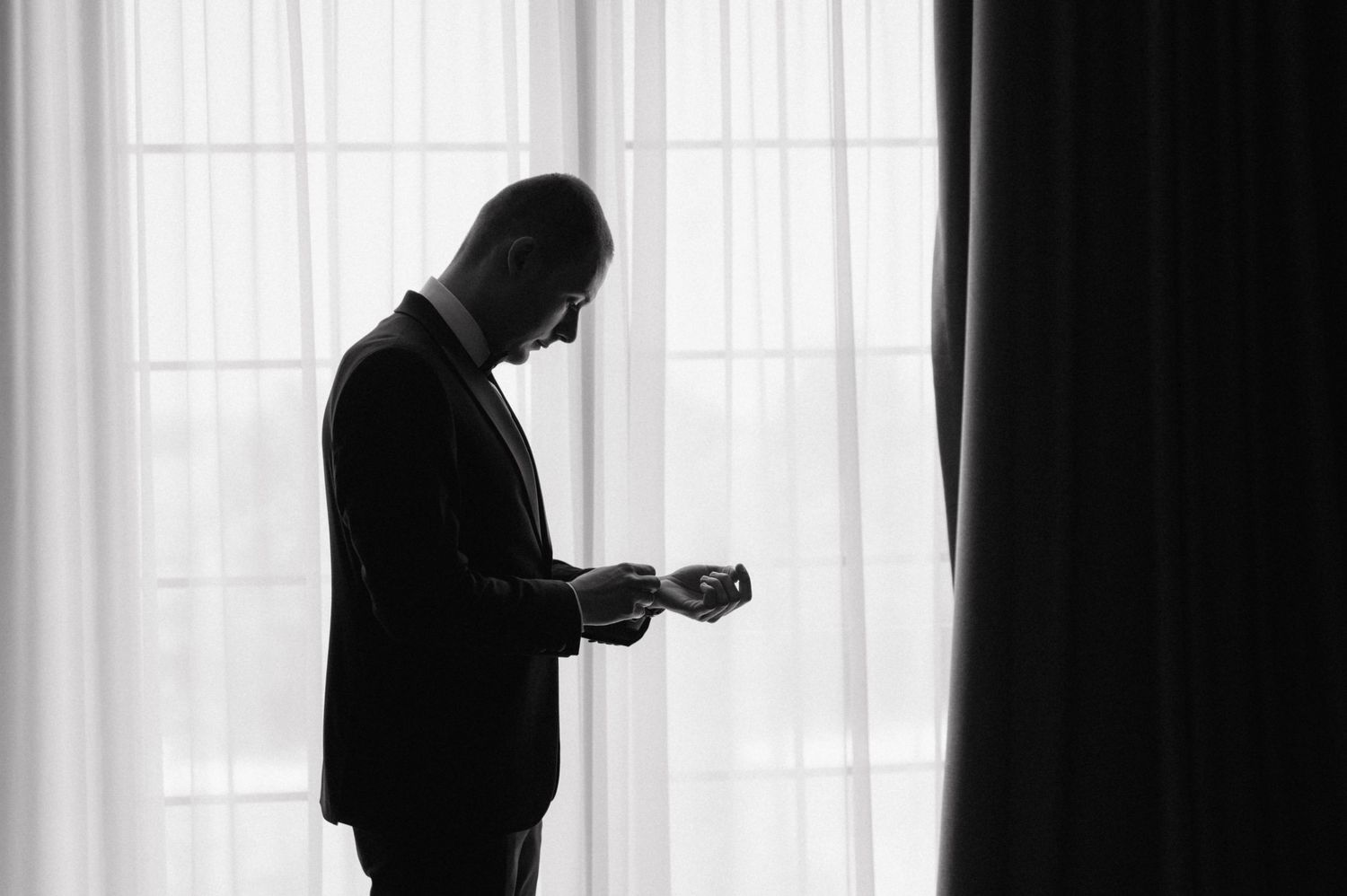 A dark silhouette stands in a bright window with vertical blinds casting dramatic shadows on the wall.