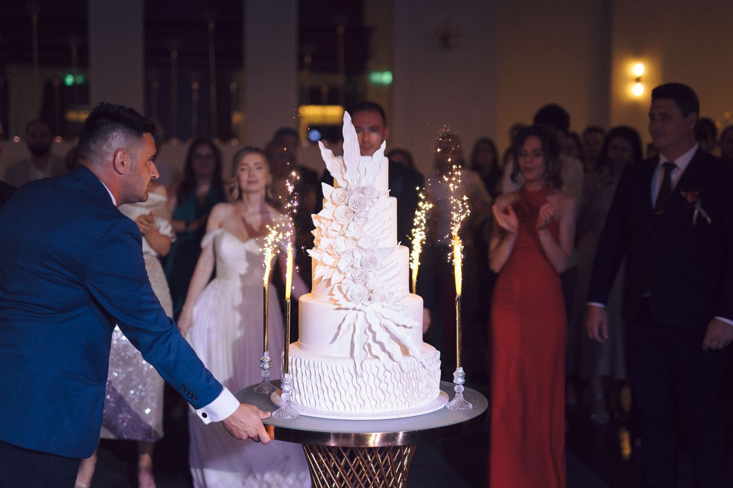 A wedding reception features multiple views of a tall white wedding cake being displayed and cut during an evening celebration.