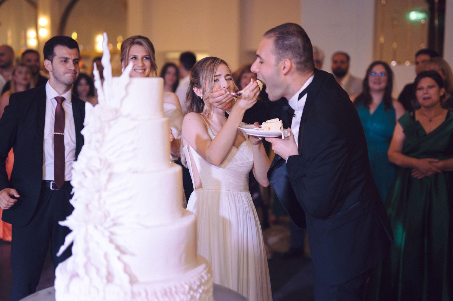 A couple feeds each other wedding cake during their reception while guests watch and smile in the background.