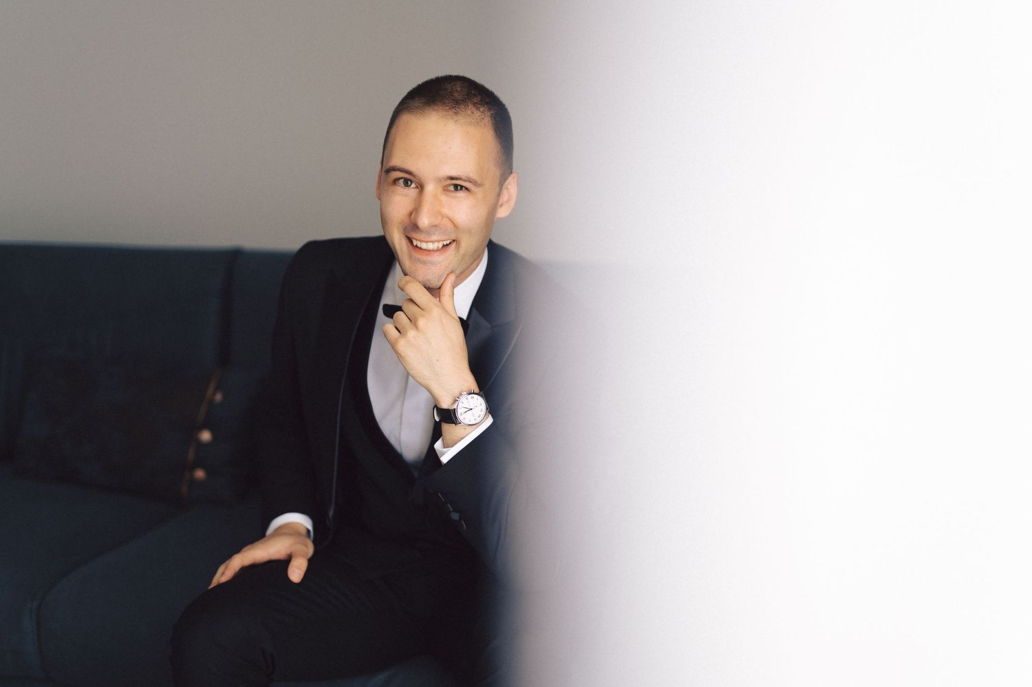 A person in a dark business suit sits on a navy couch with hand resting thoughtfully on their chin.