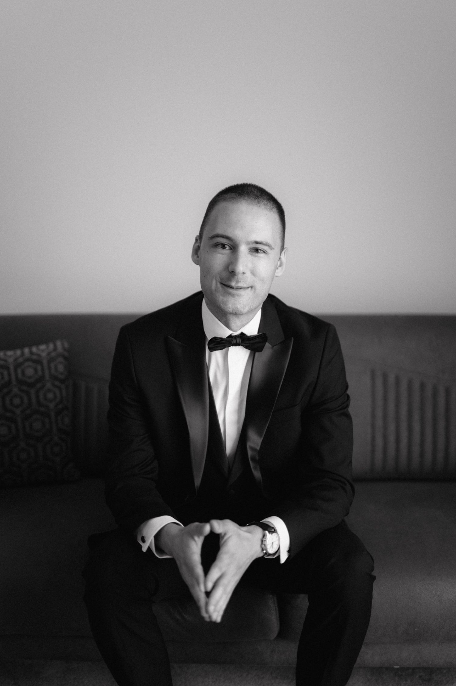 Black and white portrait of someone wearing a tuxedo with bow tie sitting on a couch looking at camera.