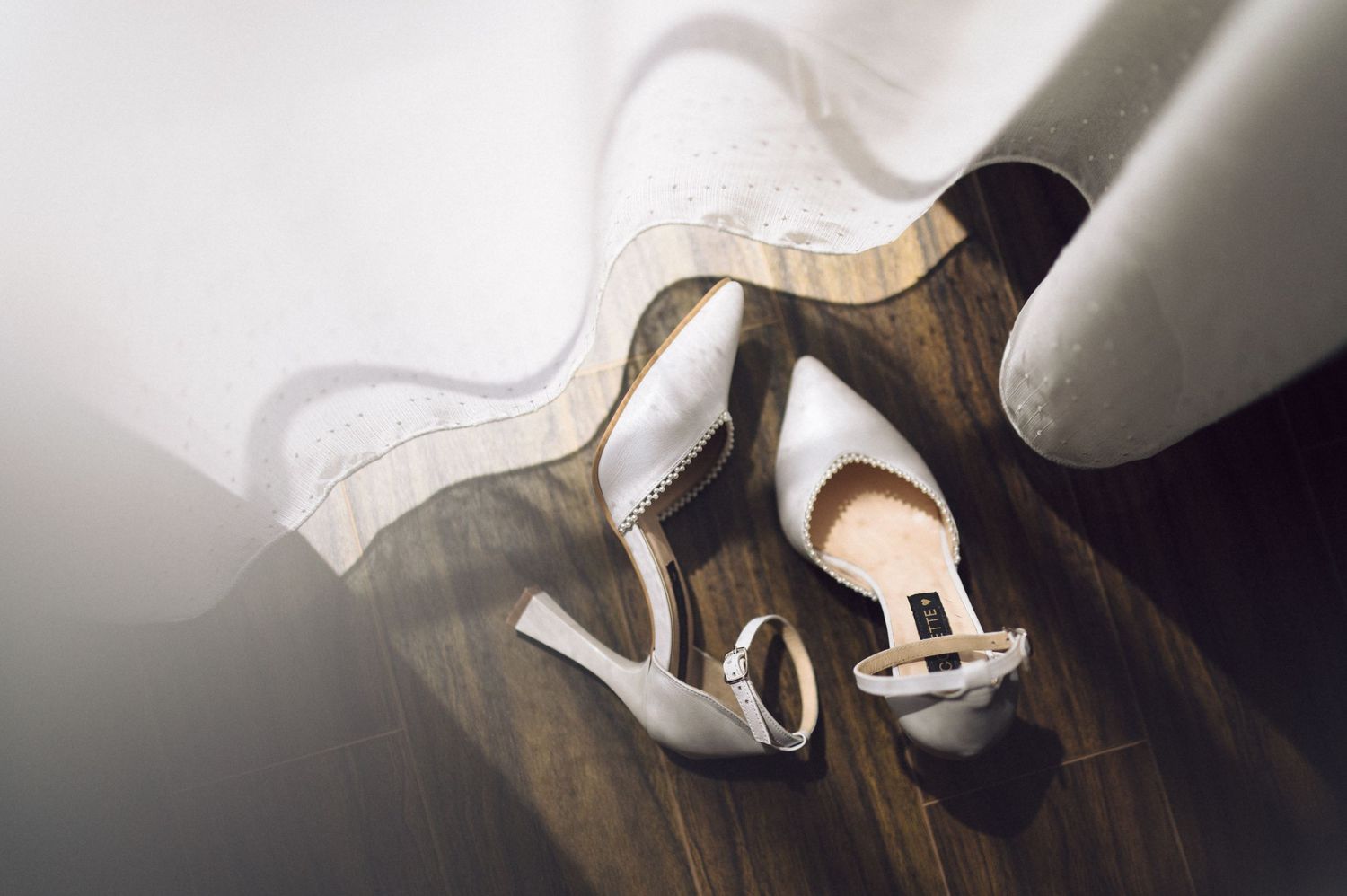 Elegant silver stiletto wedding shoes with ankle straps lying on dark wooden floor near window light.