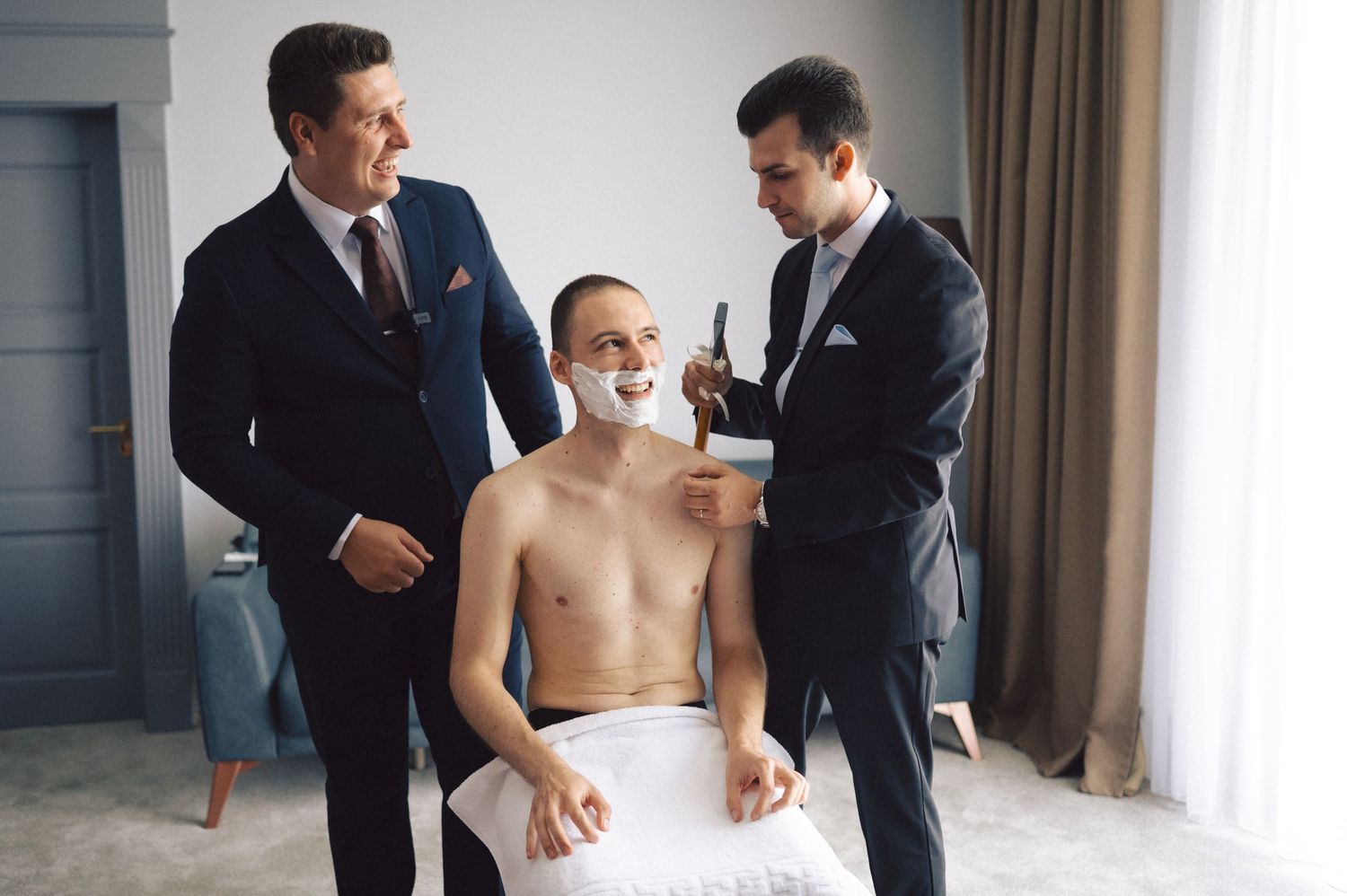 A group of well-dressed men help another man put on formal attire in a hotel room.