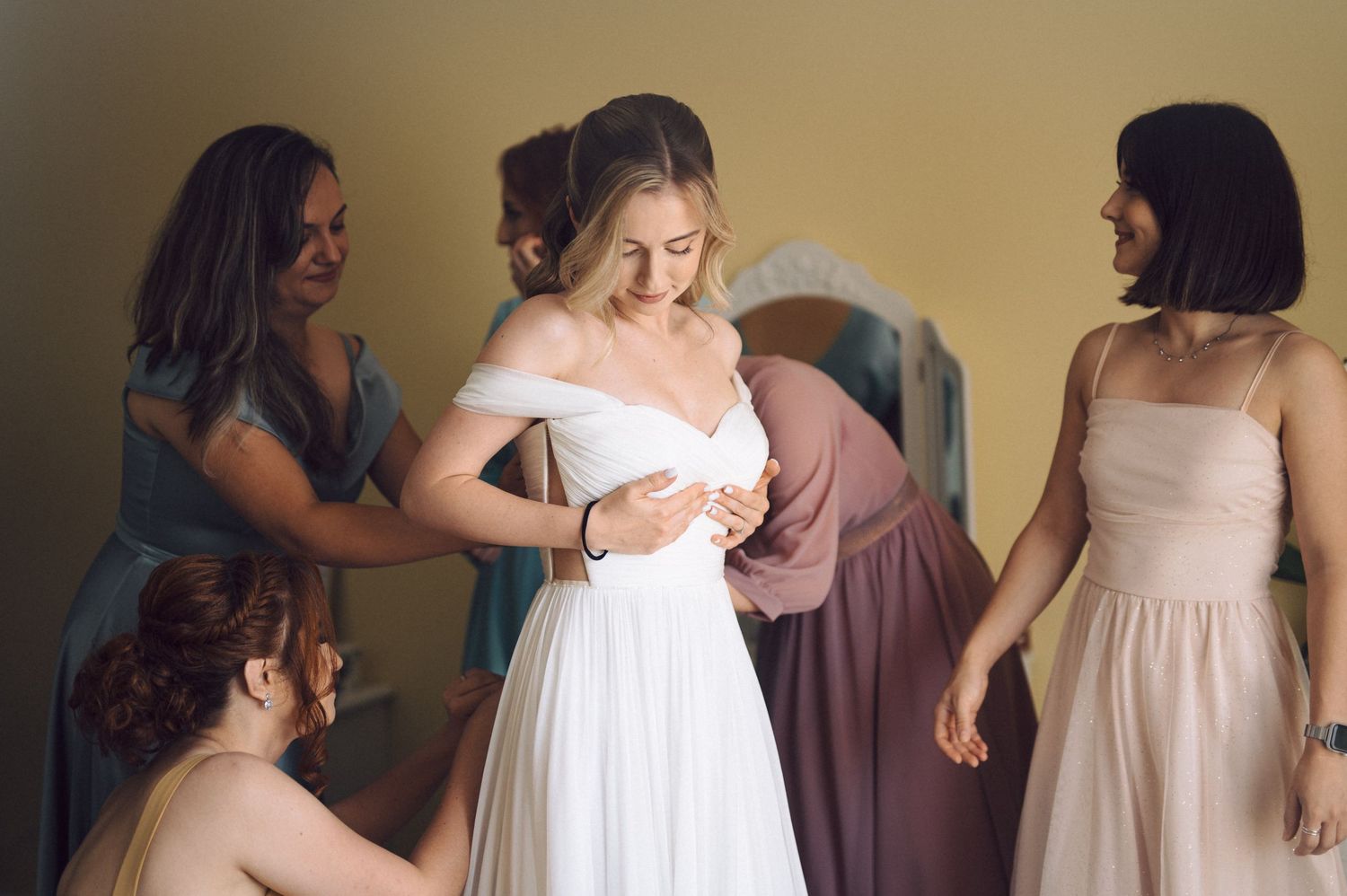 Several women assist adjusting the bride's white wedding dress in a preparation room.