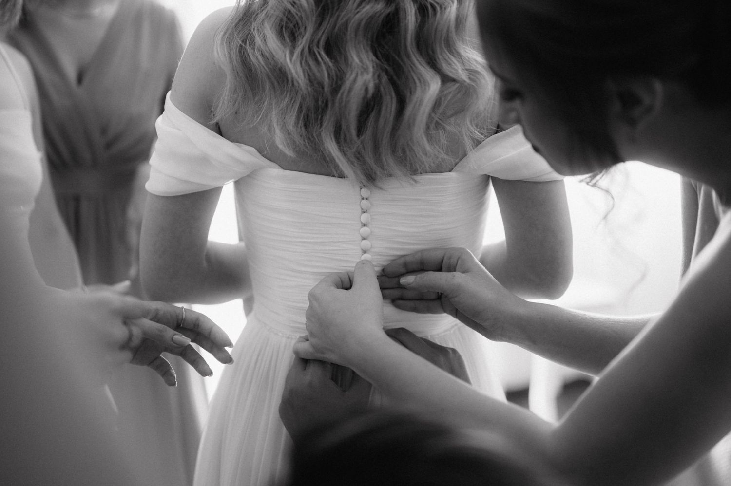 Close up black and white photo of people helping button up a delicate white wedding dress.