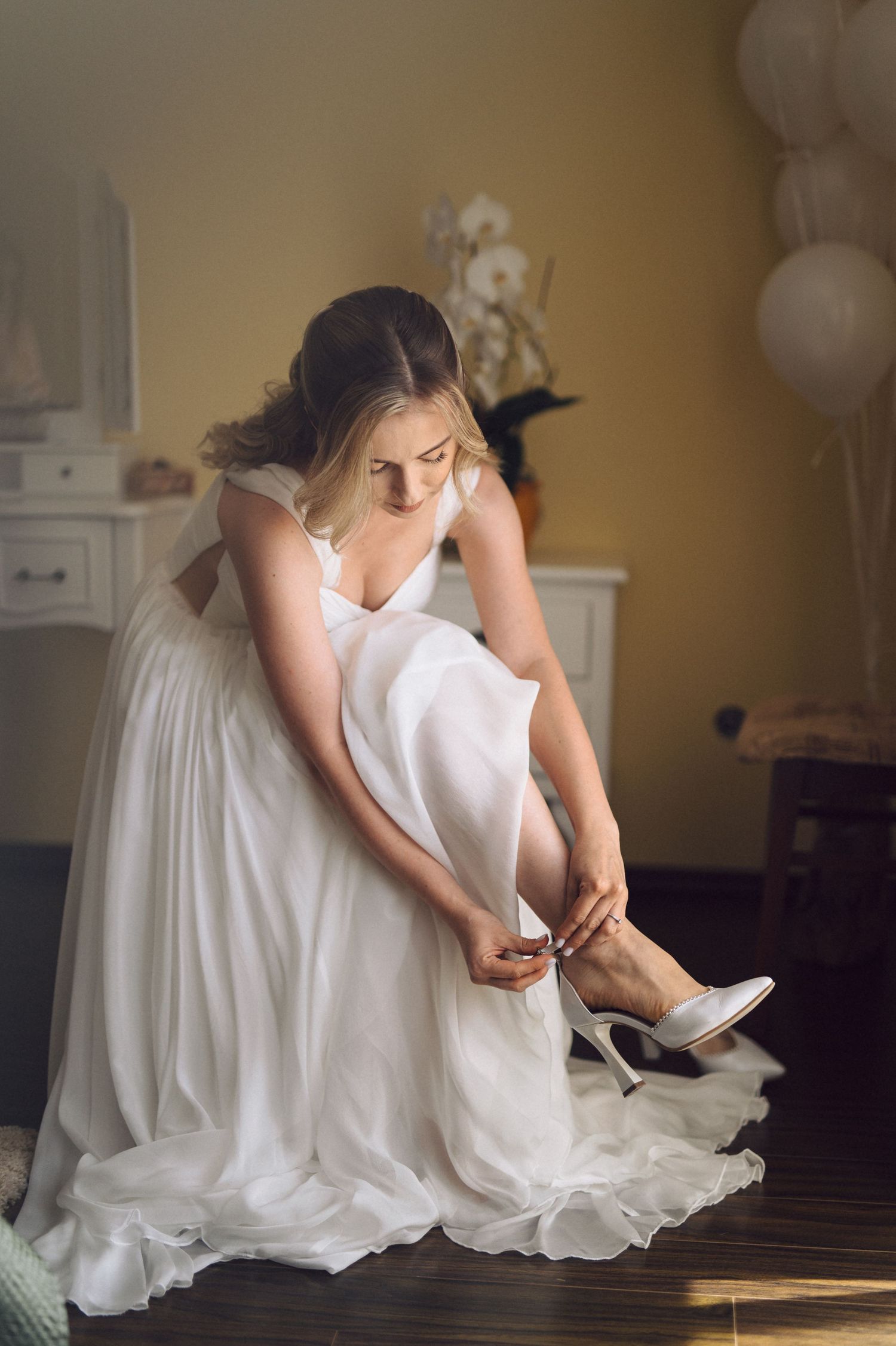 A bride in a flowing white dress fastens her shoe while sitting in a room decorated with balloons and white furniture.