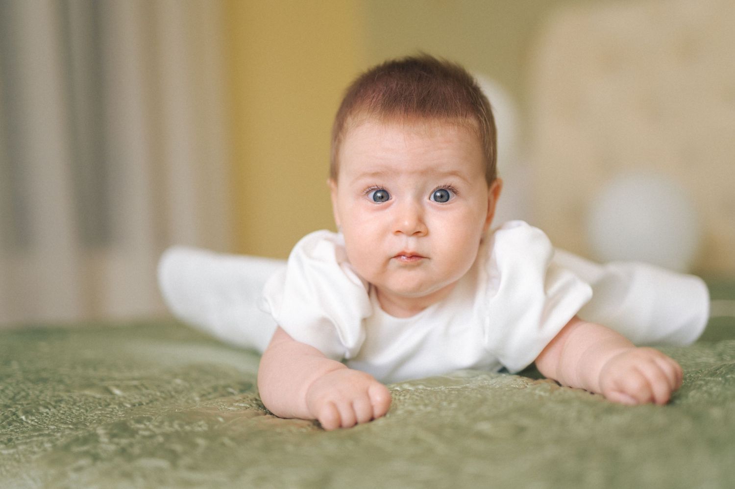 A baby wearing a white onesie lies on a soft green carpet while looking up at the camera.