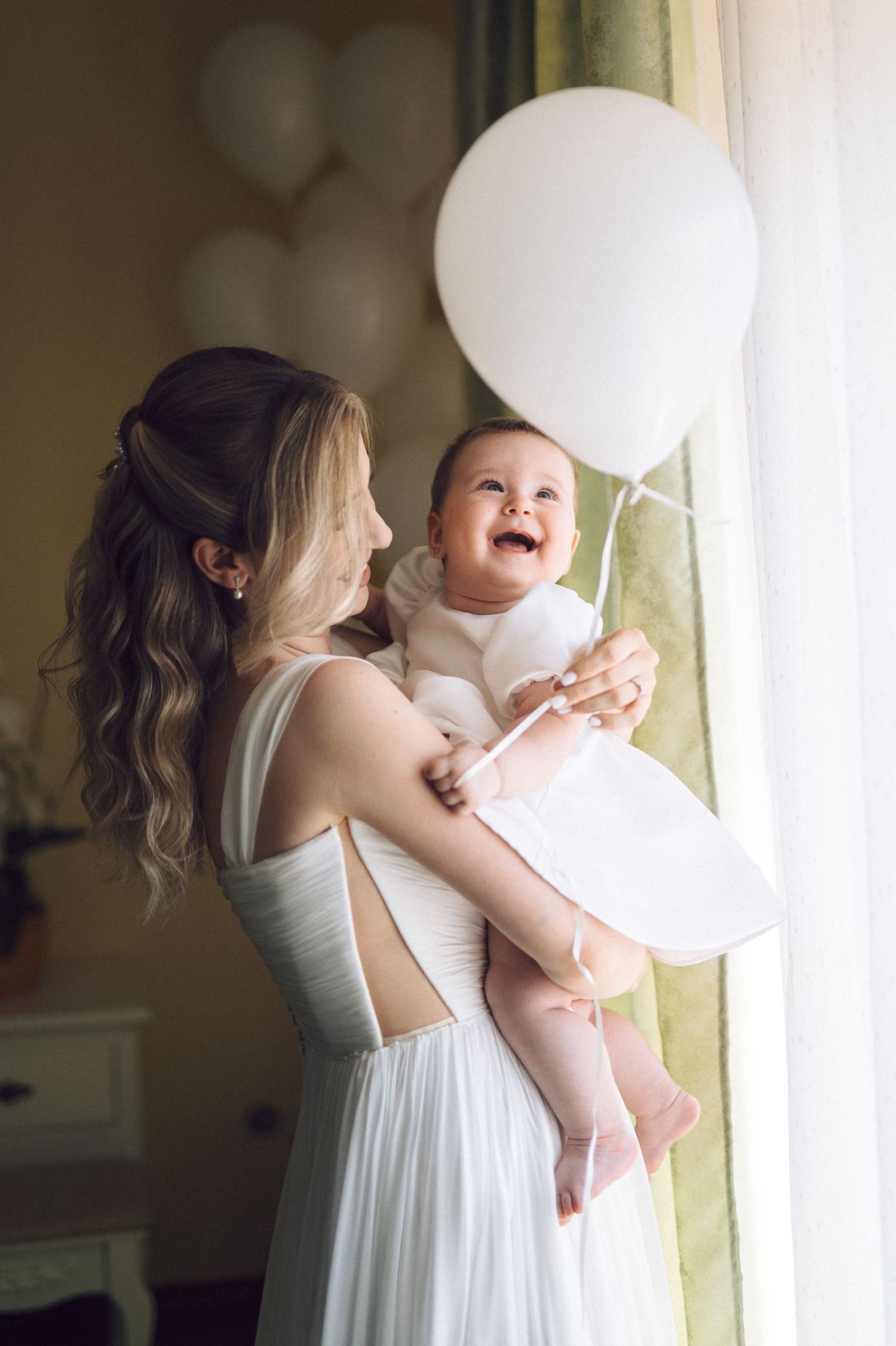 A tender moment by a window as a baby in white laughs joyfully while being held near a white balloon.