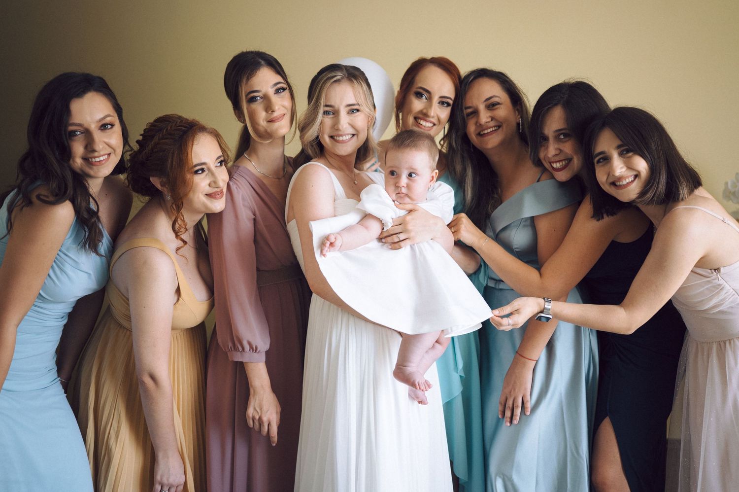 A group in pastel-colored dresses gather together smiling while holding a baby in a white dress during a celebration.