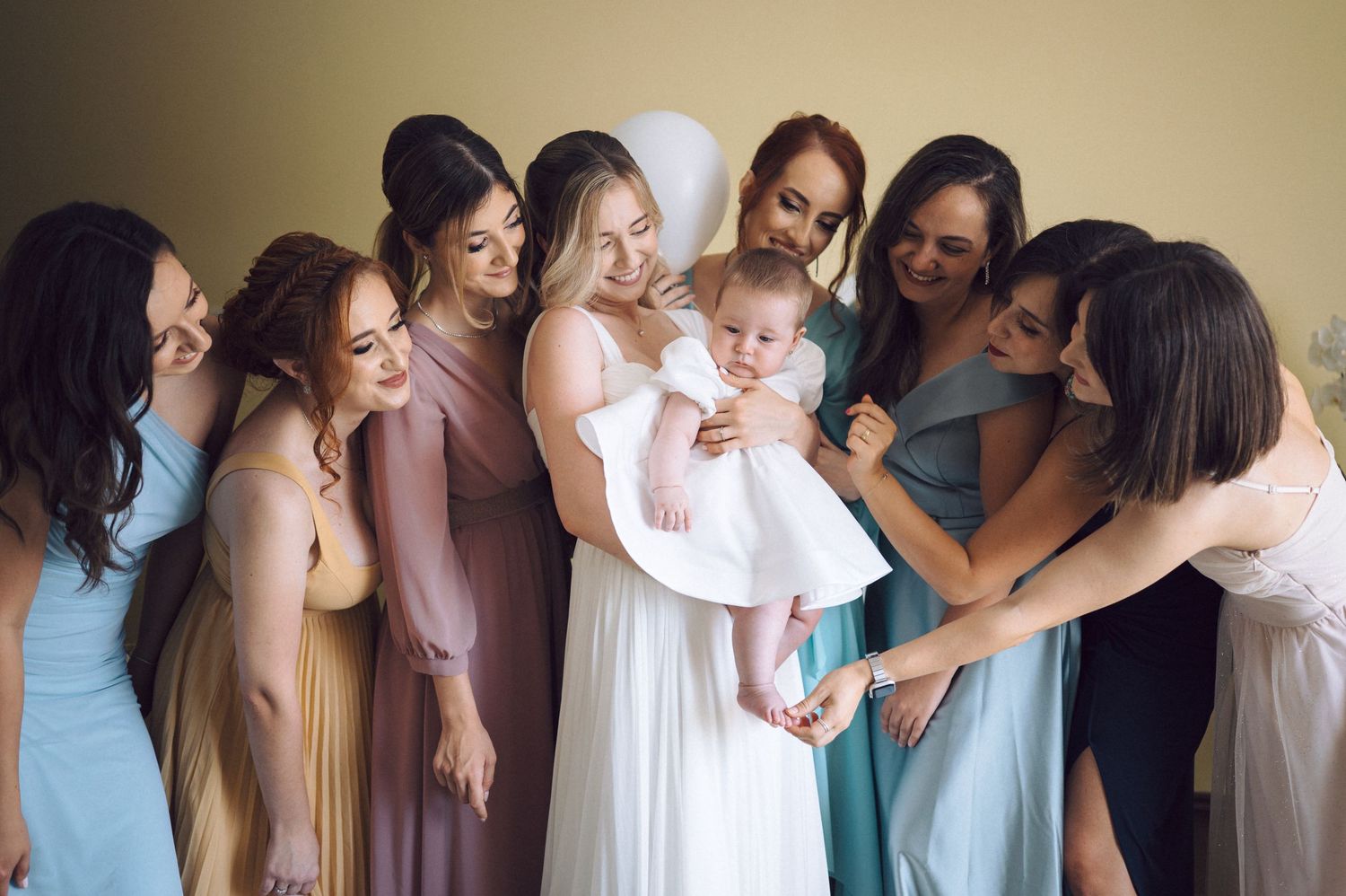 A group in pastel bridesmaid dresses gathers around a baby wearing a white dress during a wedding celebration.