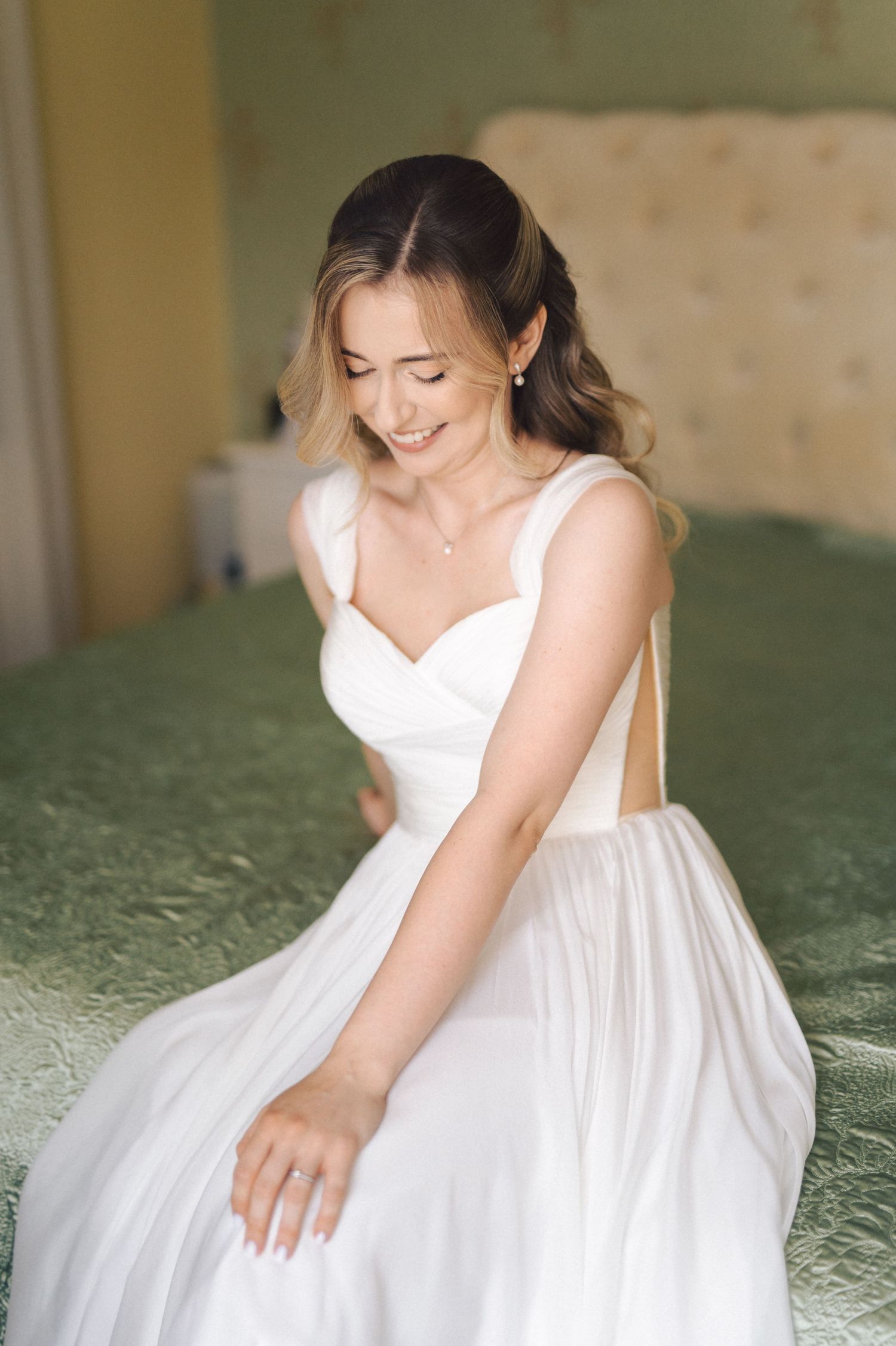 Bride in flowing white dress smiles while sitting on green upholstered furniture during wedding preparations.