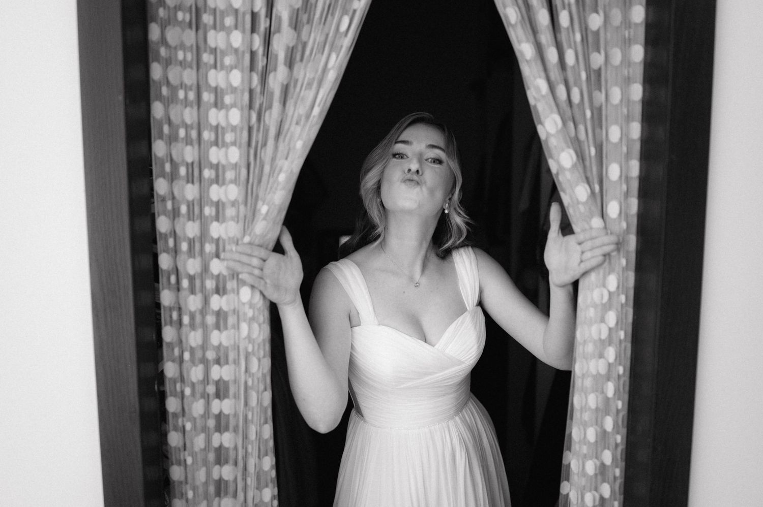 Black and white photo of a person in a white dress standing between curtains with an expressive pose.