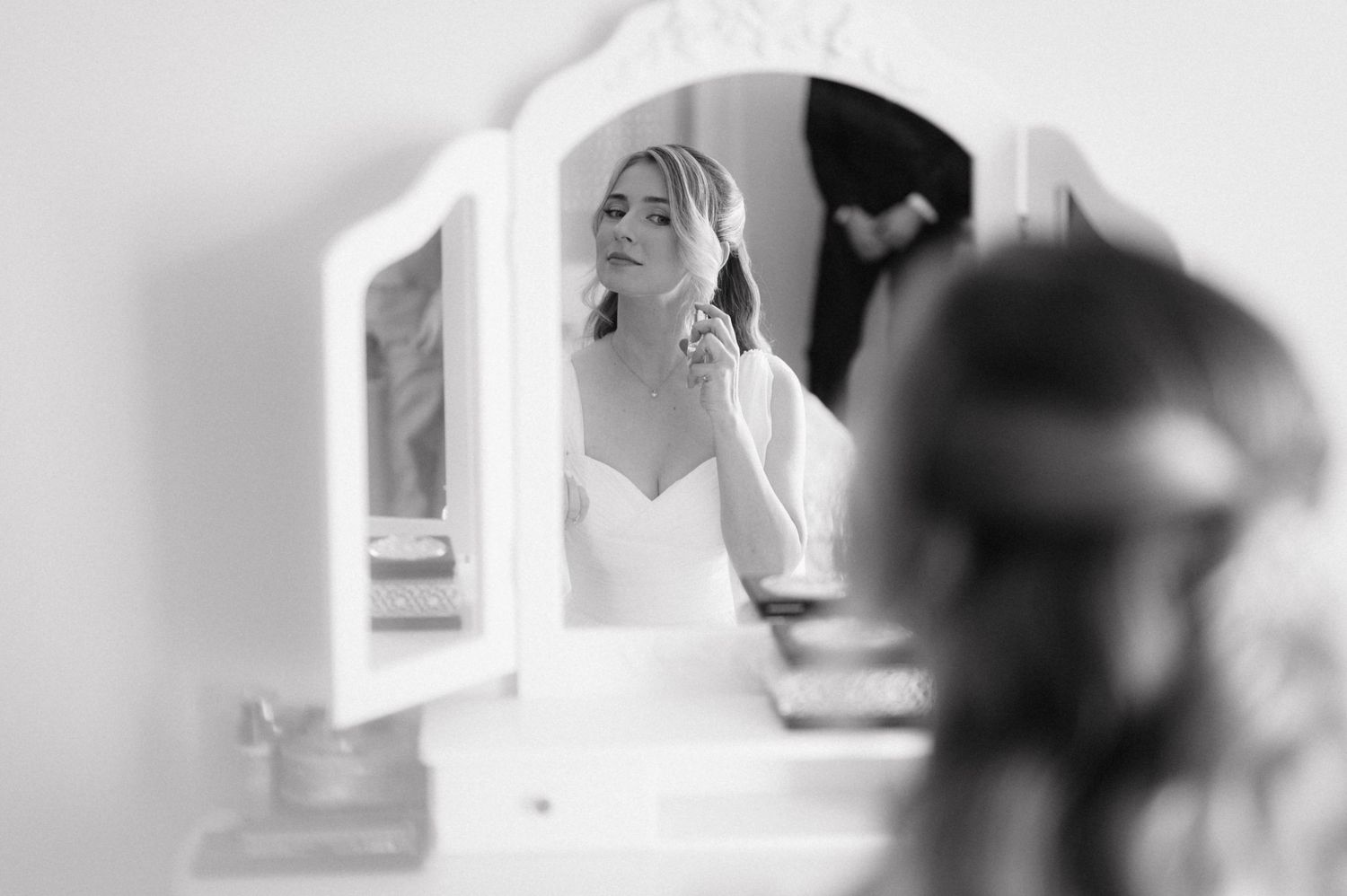 Black and white wedding photography shows bride in white dress reflected in mirror during preparation.