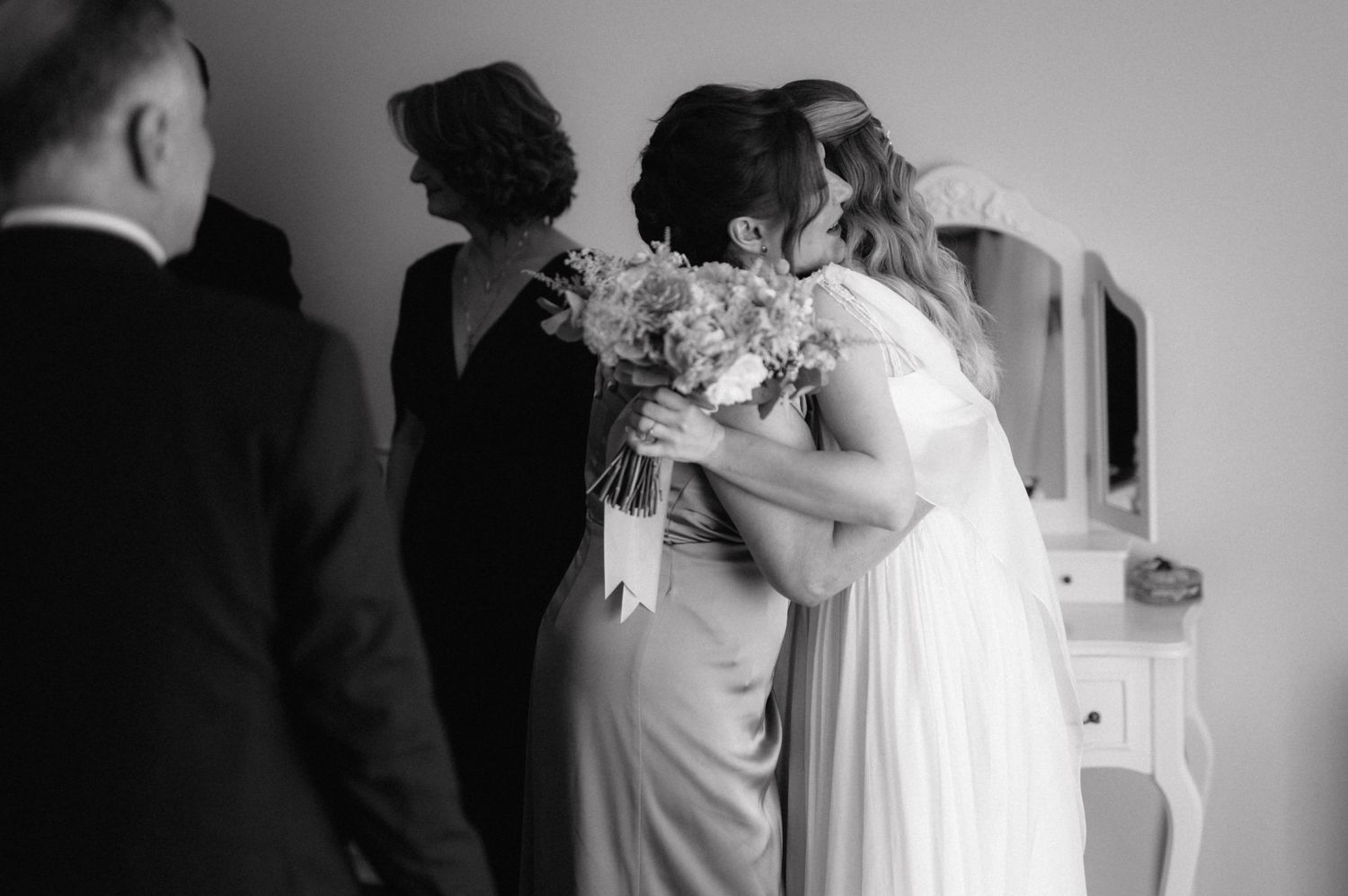 Black and white candid moment at a wedding showing bridal party members embracing with bouquet of flowers.