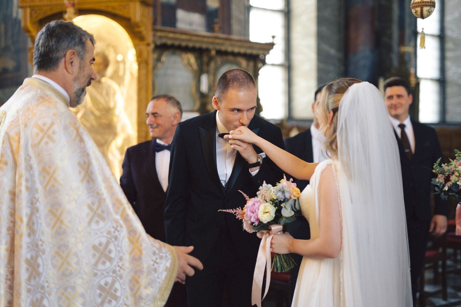 Couple exchanges vows during ornate church wedding ceremony with clergy in white vestments and guests looking on.