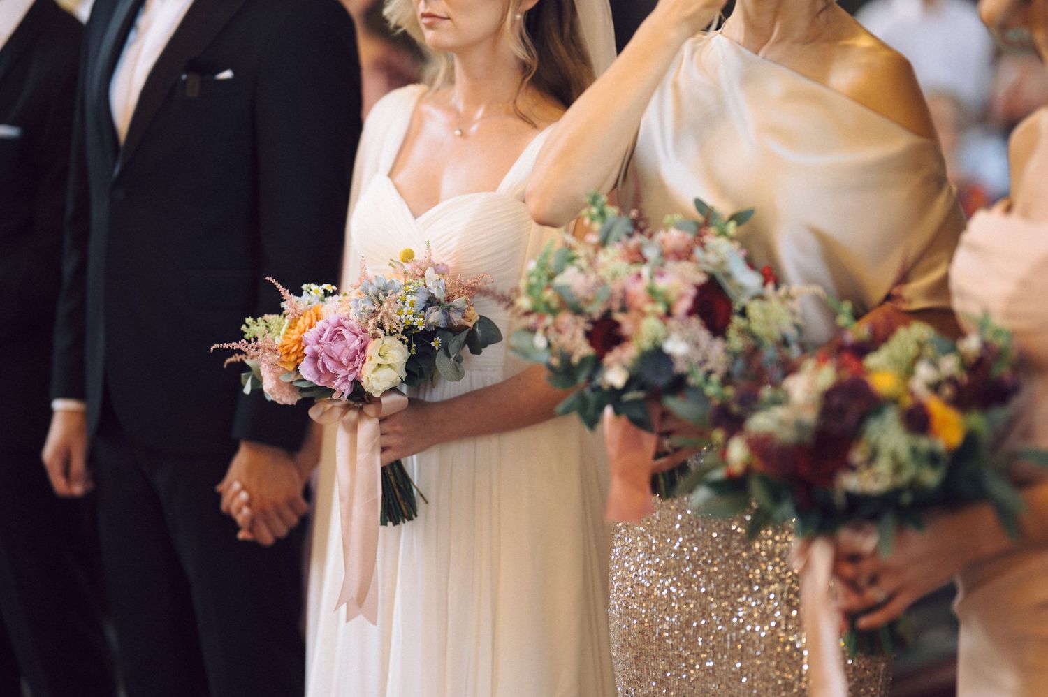 Bridesmaids in champagne colored gowns holding pastel bouquets line up during wedding ceremony.