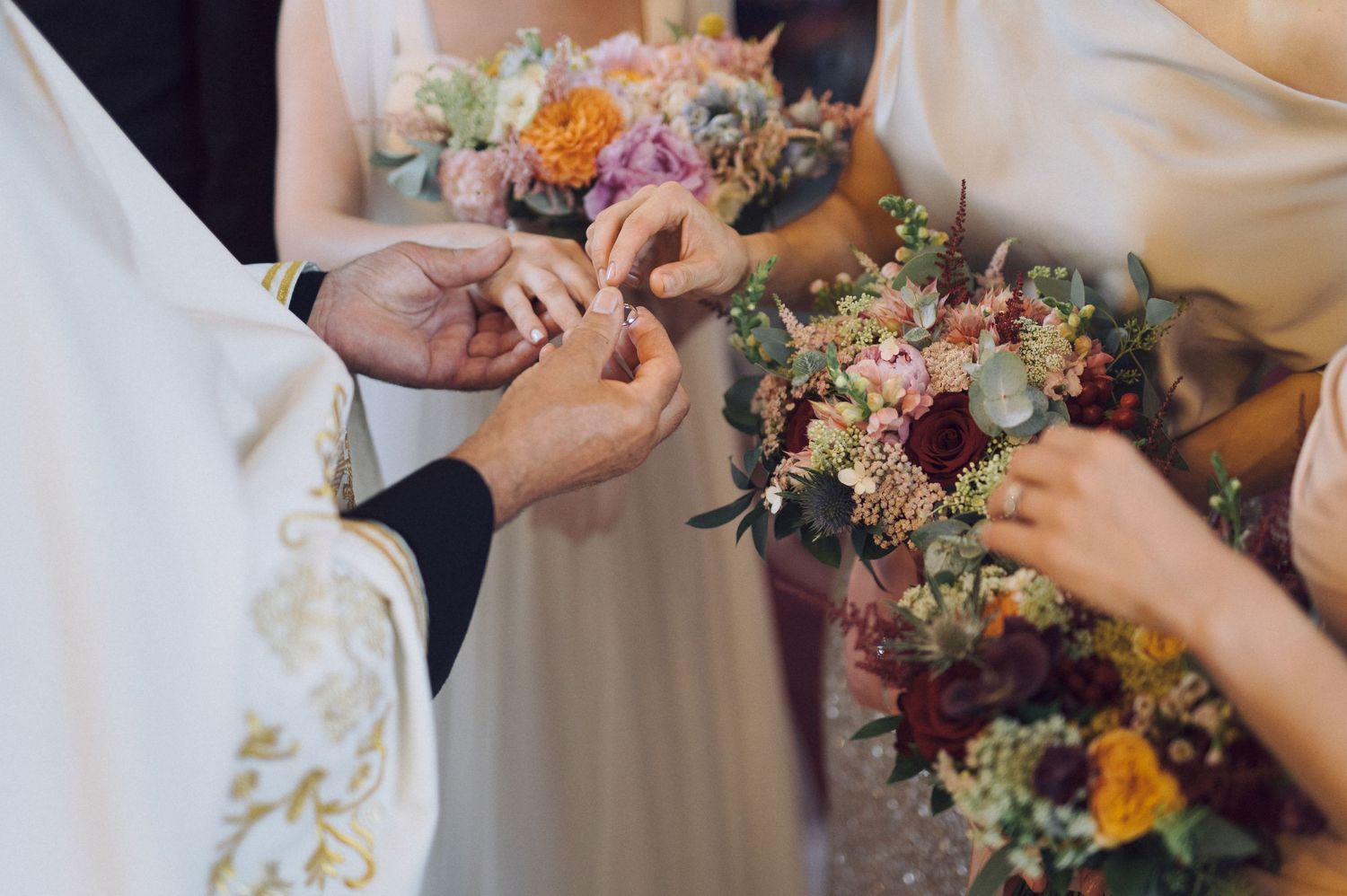 A close-up shot of hands exchanging rings during a wedding ceremony, with colorful bouquets of orange and pink flowers.