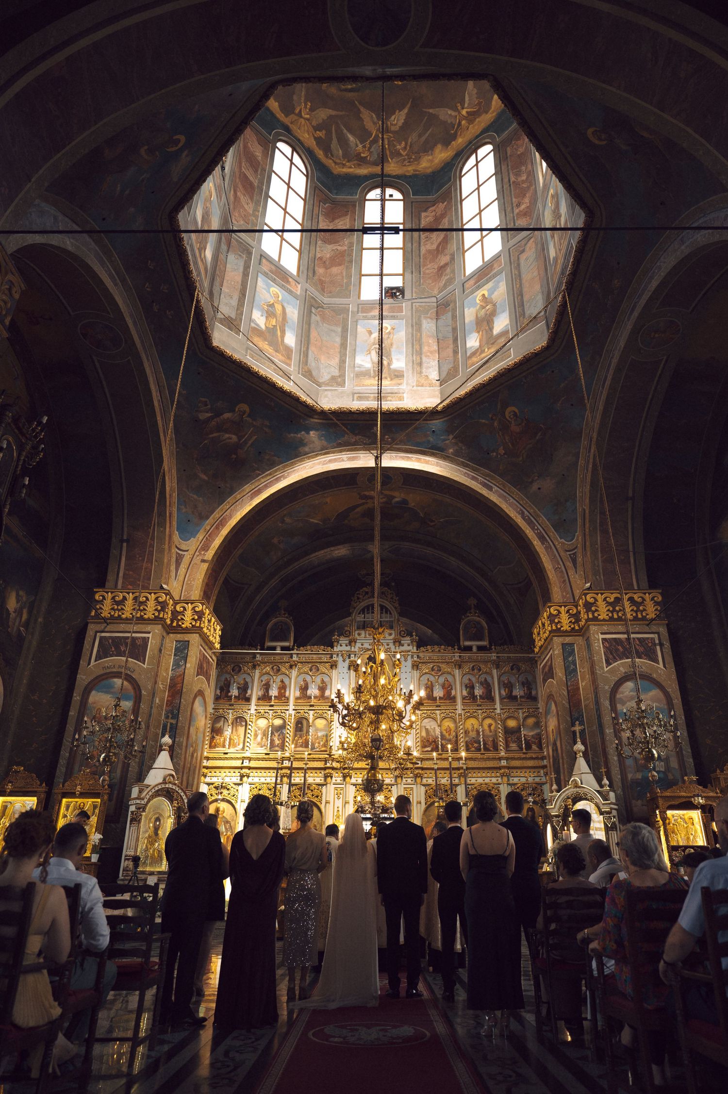 A solemn church service takes place beneath a dramatic domed ceiling with ornate icons and golden Orthodox iconostasis.