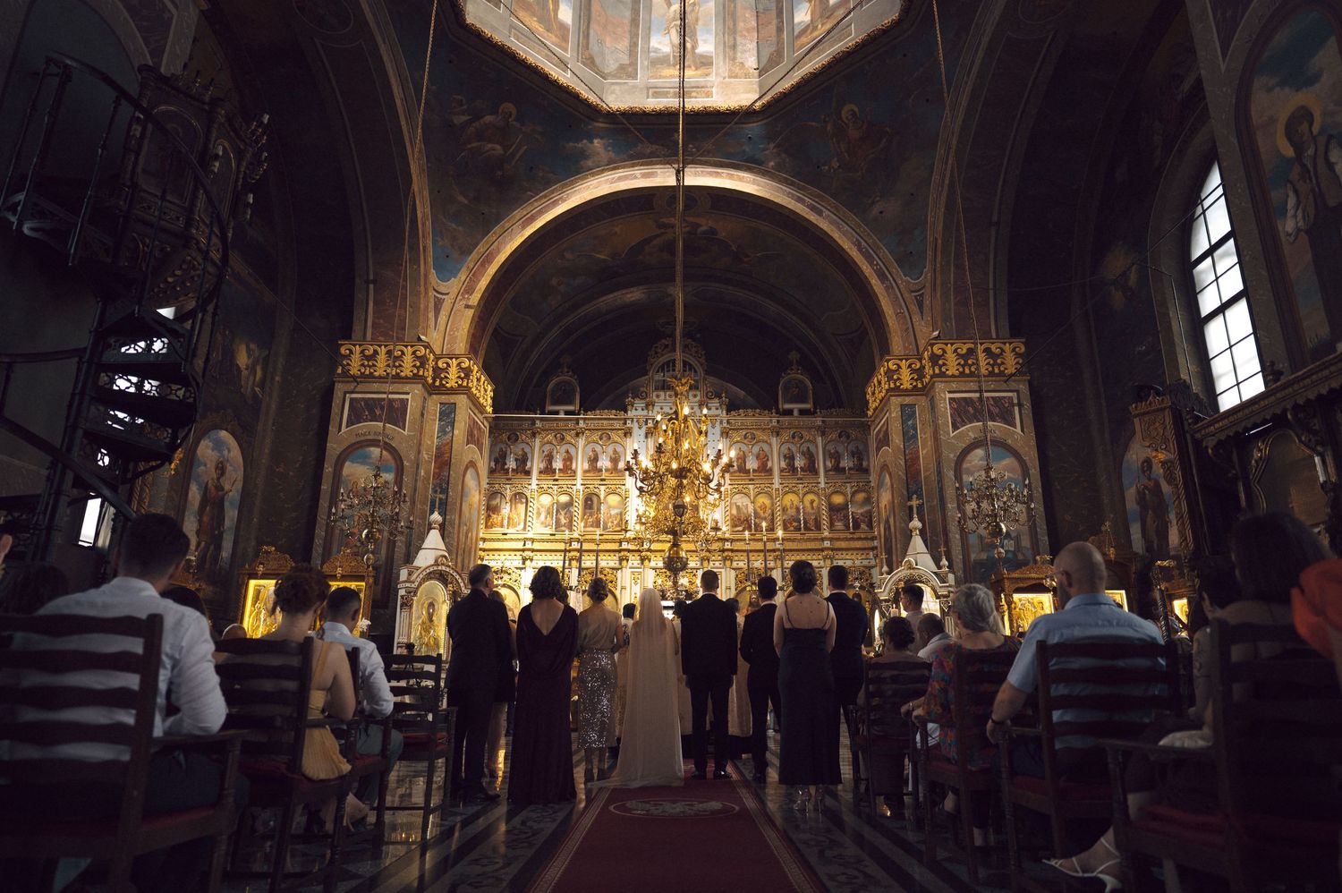 Interior view of an Orthodox church showing parishioners standing before an ornate golden iconostasis during a service.