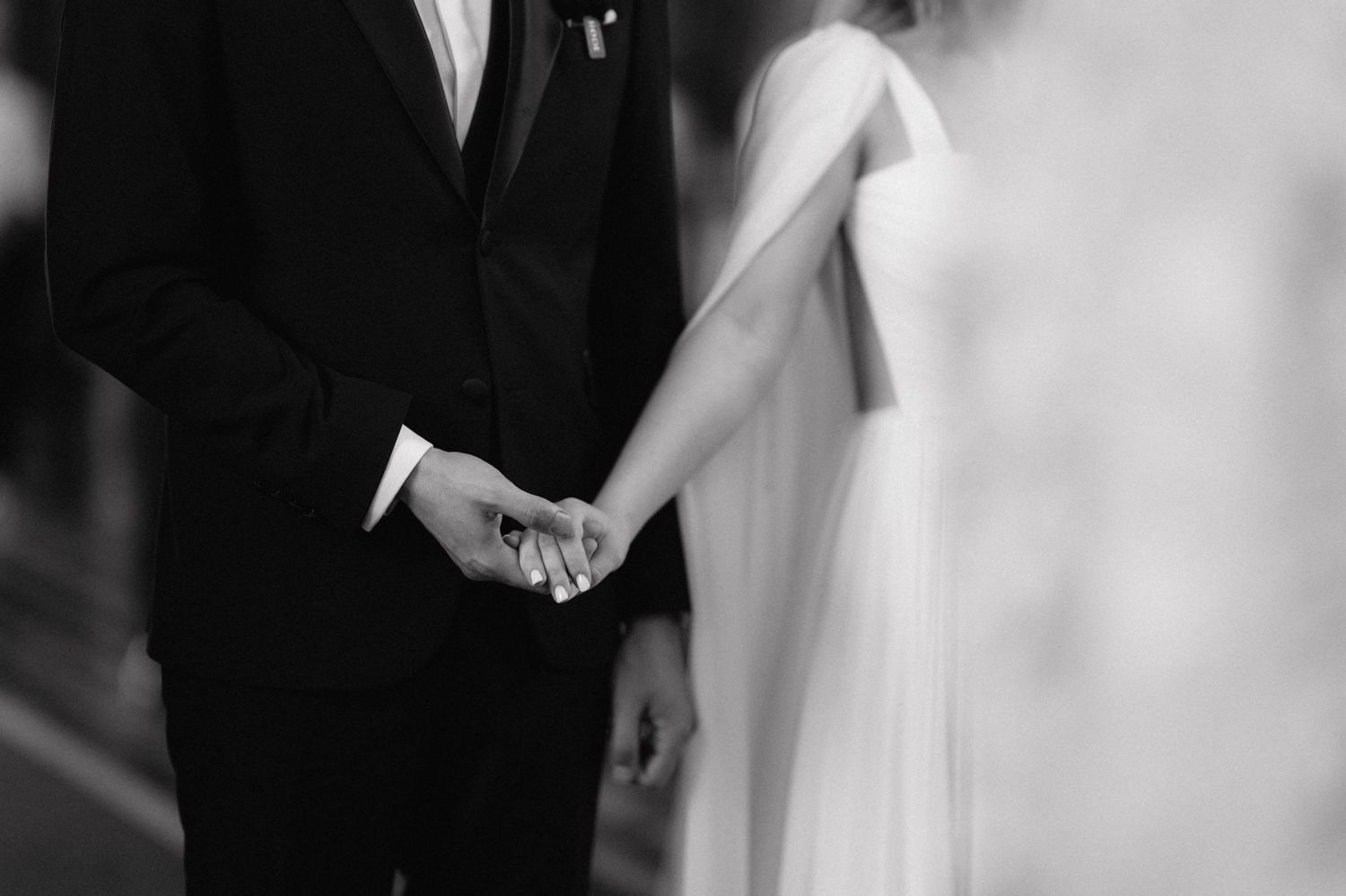 A black and white close-up photo shows a couple holding hands at their wedding ceremony.