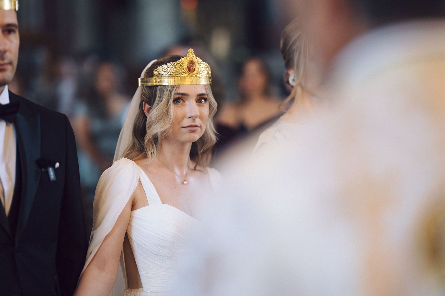 An elegant bride wearing a white one-shoulder gown and ornate gold crown headpiece during a wedding ceremony.