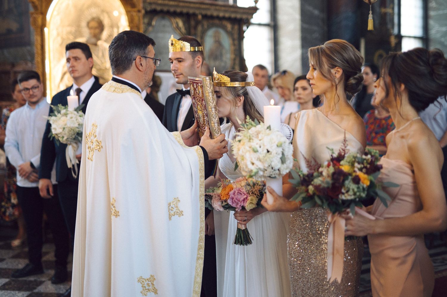 A priest leads a wedding ceremony in an ornate church while attendants holding bouquets stand in formal attire.