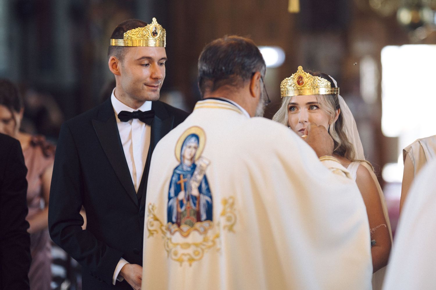 Religious ceremony participants wearing golden crowns stand together during an ornate Orthodox Christian wedding service.
