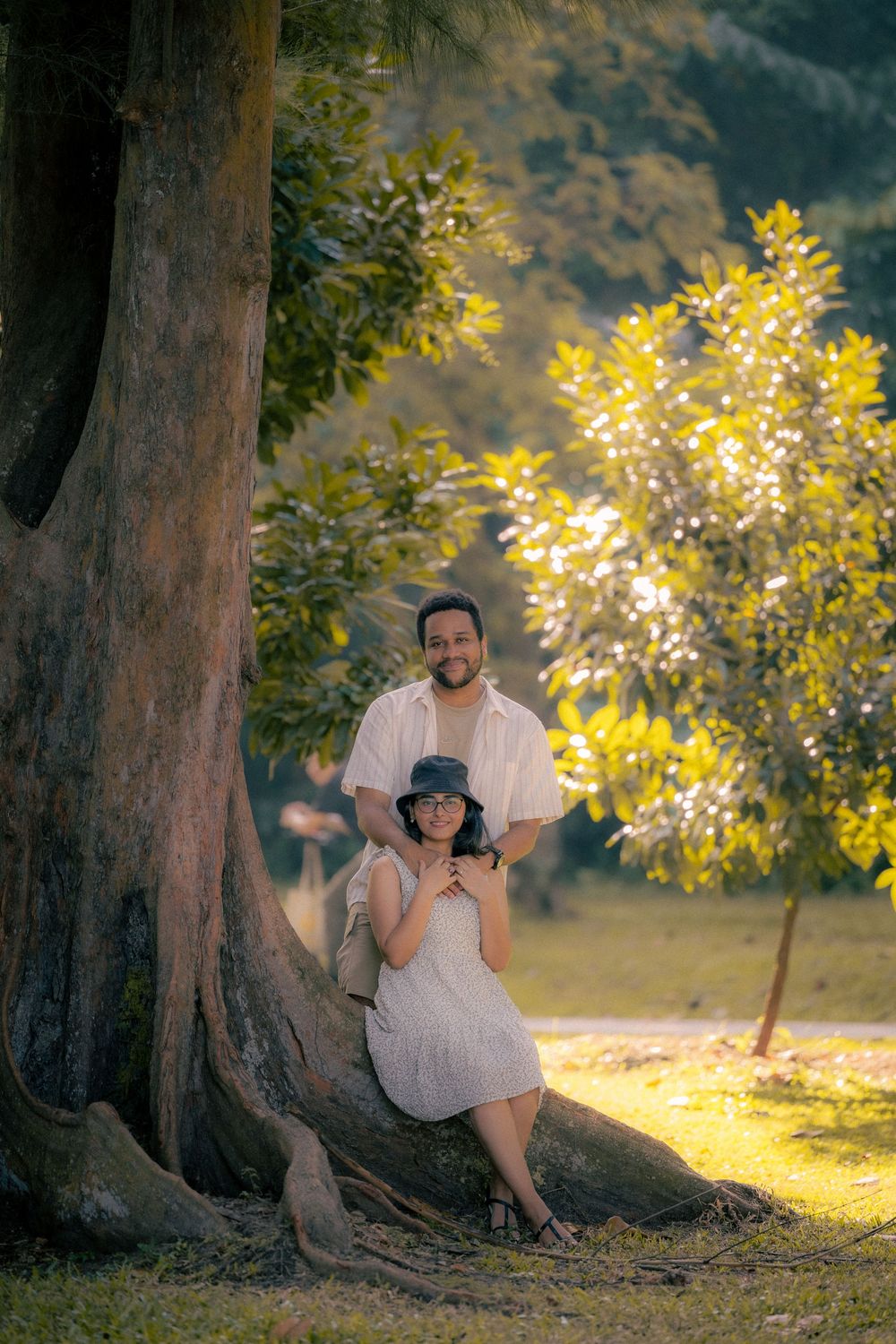 A couple sits together at the base of large trees in a park during golden hour sunlight.