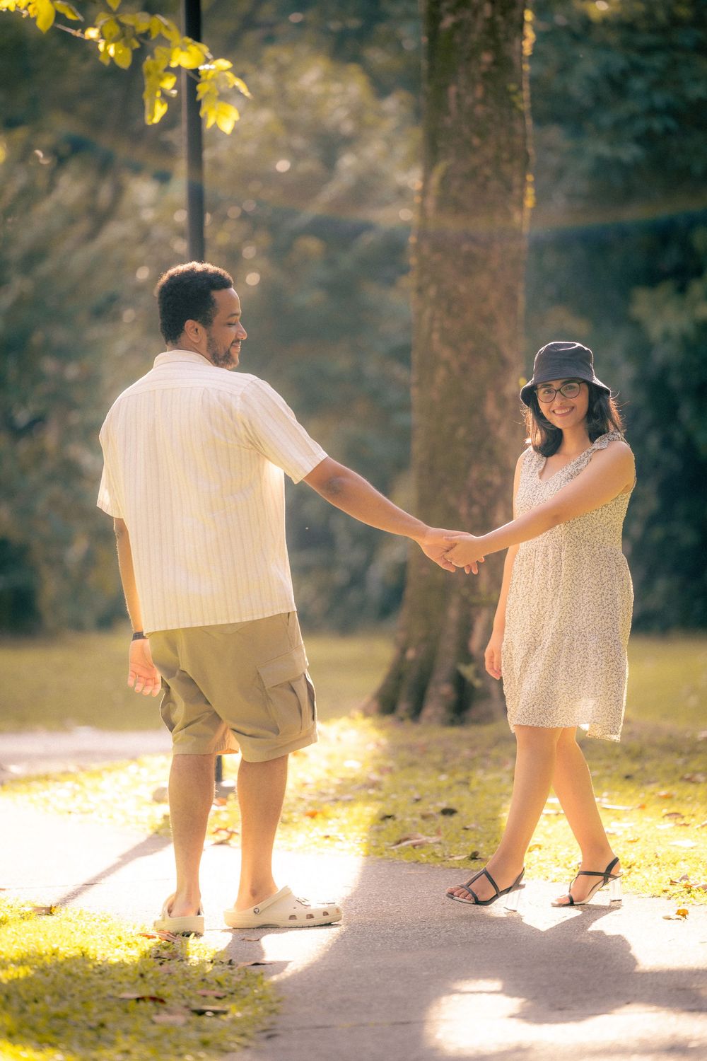 A couple holding hands on a sunlit path in a park during golden hour, surrounded by trees and dappled sunlight.