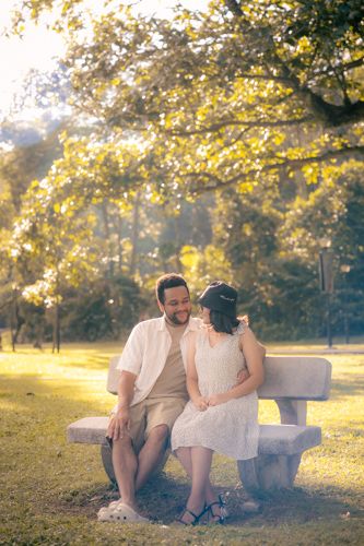 A couple sits close together on a stone bench in a sunlit park with golden autumn foliage in the background.