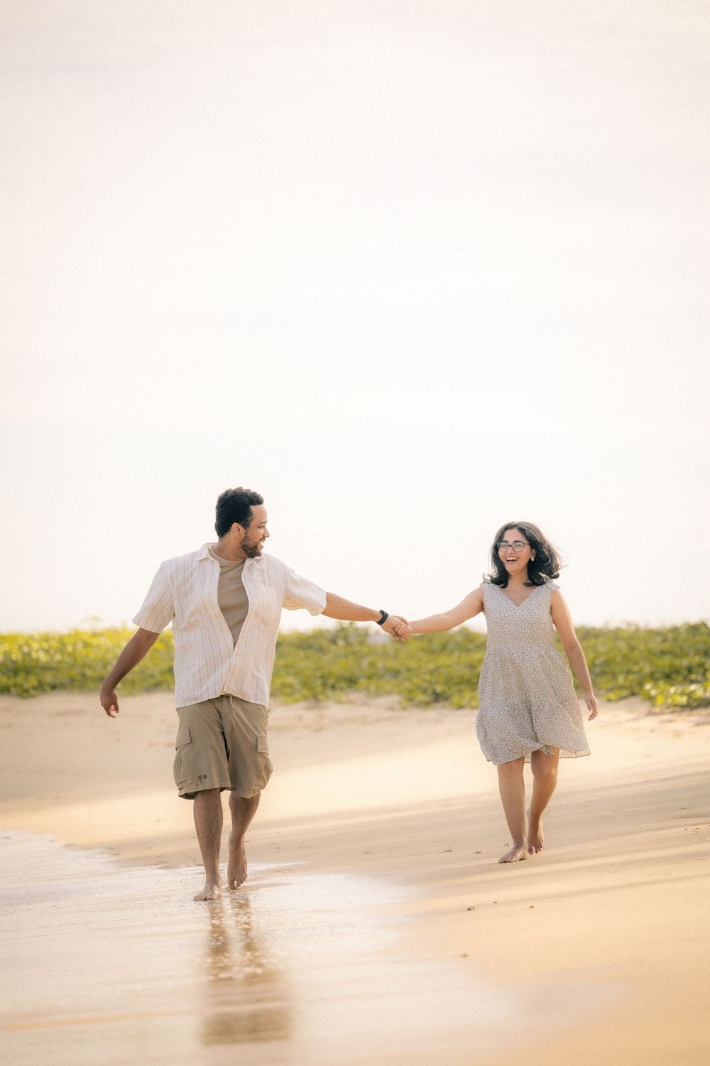 Two people hold hands while walking barefoot on a sunny beach with a golden field in the background.
