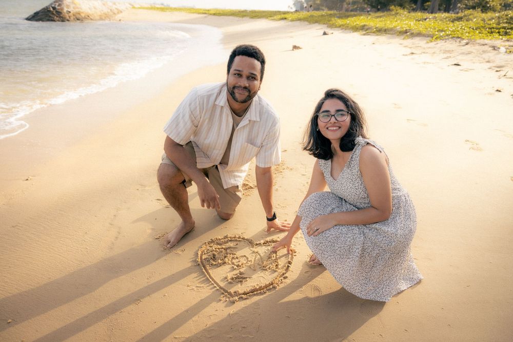 A couple kneels on a sunny beach drawing a heart in the sand at sunset.