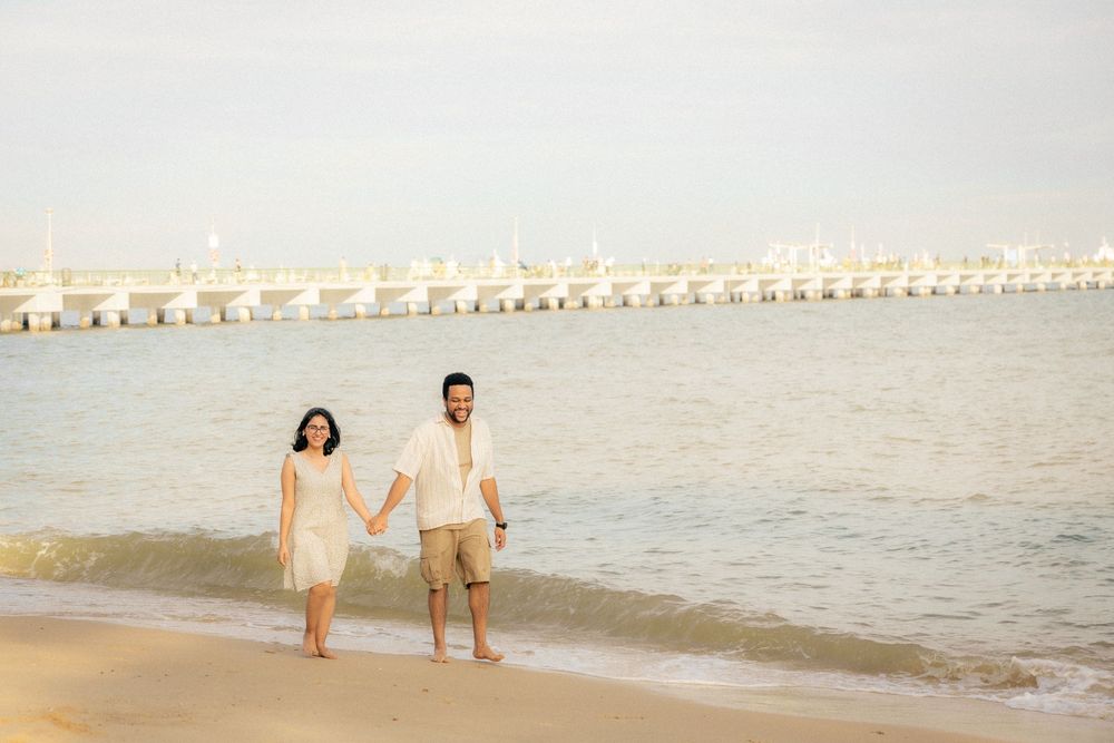 A couple walks together along a peaceful beach during golden hour with a long pier stretching into the water behind them.