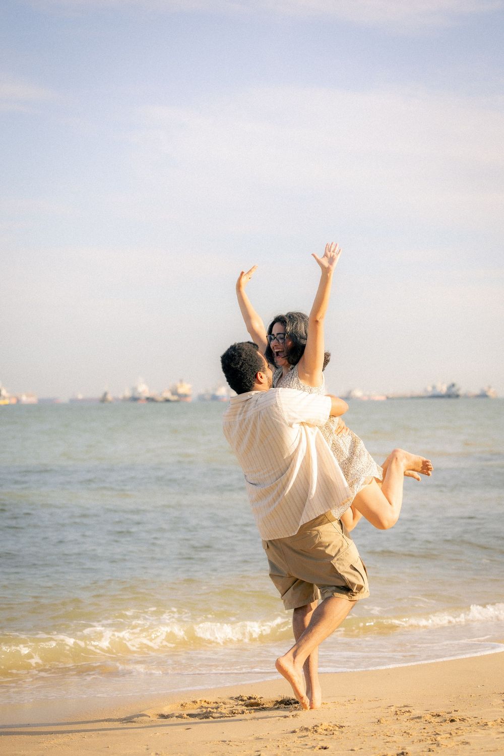 Two people in white clothes jumping joyfully together on a beach with ocean and ships in the background.